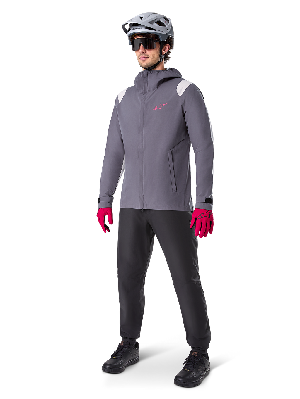 Modèle masculin portant la veste de pluie Alpinestars A-Dura en Titanium Foncé, veste grise à capuche avec logo rose sur la poitrine et empiècements gris clair aux épaules, complétée par un casque de VTT gris, des lunettes de soleil, des gants rose vif et un pantalon noir pour le cyclisme tout-terrain