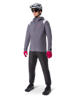 Modèle masculin portant la veste de pluie Alpinestars A-Dura en Titanium Foncé, veste grise à capuche avec logo rose sur la poitrine et empiècements gris clair aux épaules, complétée par un casque de VTT gris, des lunettes de soleil, des gants rose vif et un pantalon noir pour le cyclisme tout-terrain