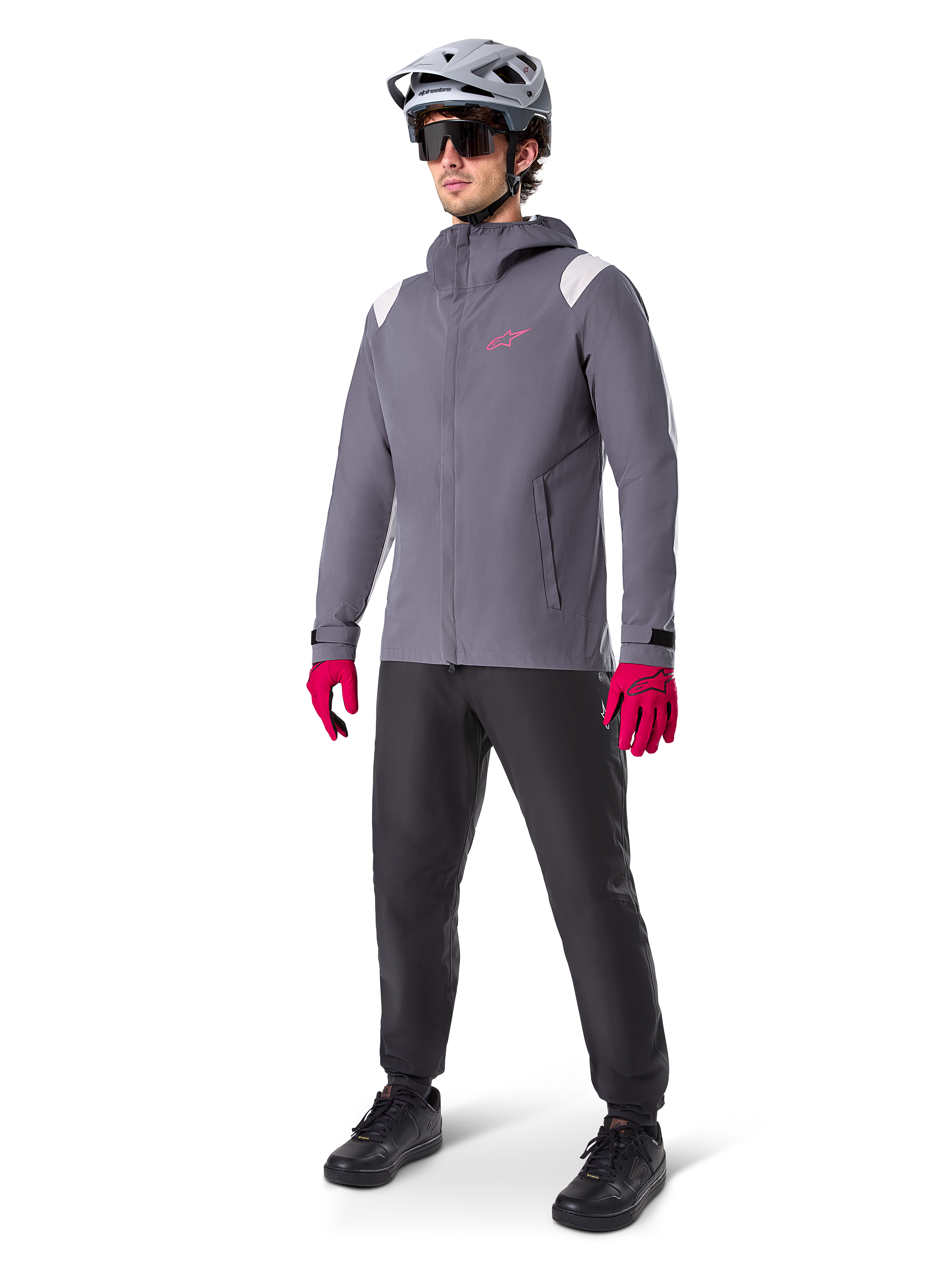 Modèle masculin portant la veste de pluie Alpinestars A-Dura en Titanium Foncé, veste grise à capuche avec logo rose sur la poitrine et empiècements gris clair aux épaules, complétée par un casque de VTT gris, des lunettes de soleil, des gants rose vif et un pantalon noir pour le cyclisme tout-terrain