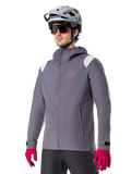 Veste de pluie Alpinestars A-Dura, veste de pluie pour VTT, gris titane foncé avec logo rose et accents blancs sur les épaules, modèle à capuche avec poches avant, portée avec des gants roses et un casque de vélo gris clair