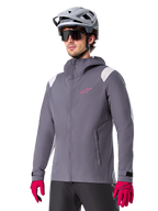 Veste de pluie Alpinestars A-Dura, veste de pluie pour VTT, gris titane foncé avec logo rose et accents blancs sur les épaules, modèle à capuche avec poches avant, portée avec des gants roses et un casque de vélo gris clair