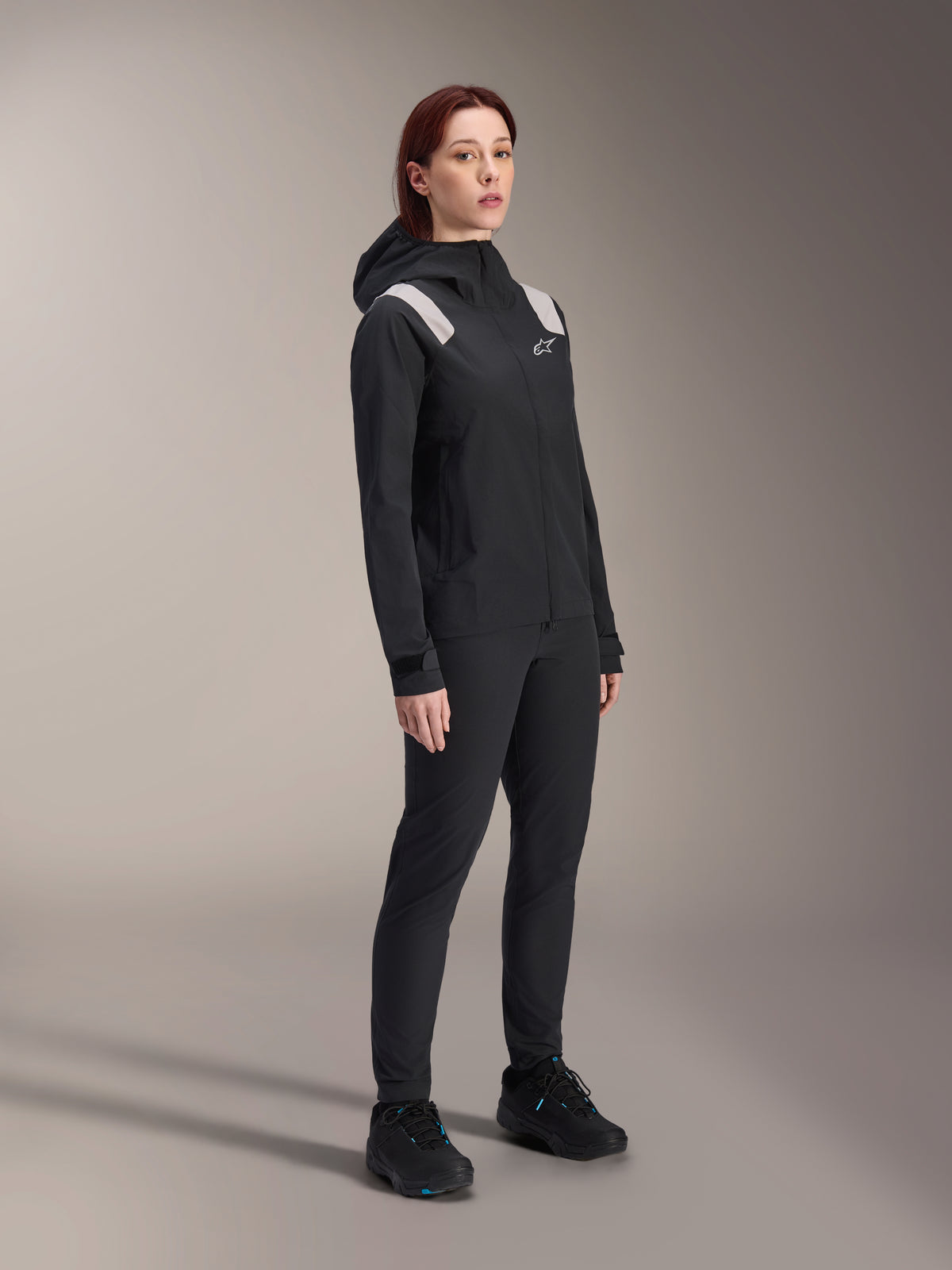 Vrouw draagt de Alpinestars Stella A-Dura Regenjas in het zwart, 2,5-laagse waterdichte MTB-shell met lichtgrijze schouderaccenten, bijpassende zwarte broek en fietsschoenen, studioportret voor performance fietskleding