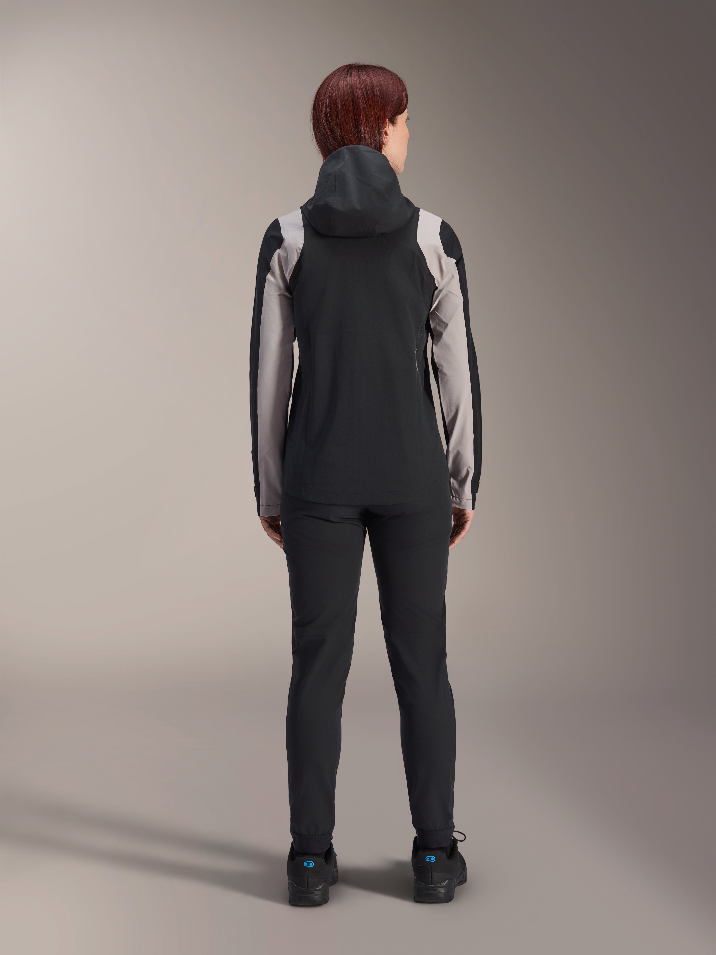 Cycliste vue de dos portant la veste de pluie Alpinestars Stella A-Dura, noire avec des empiècements gris clair sur les manches et les épaules, capuche relevée, portée avec un pantalon noir et des chaussures de cyclisme, équipement technique de pluie pour le VTT