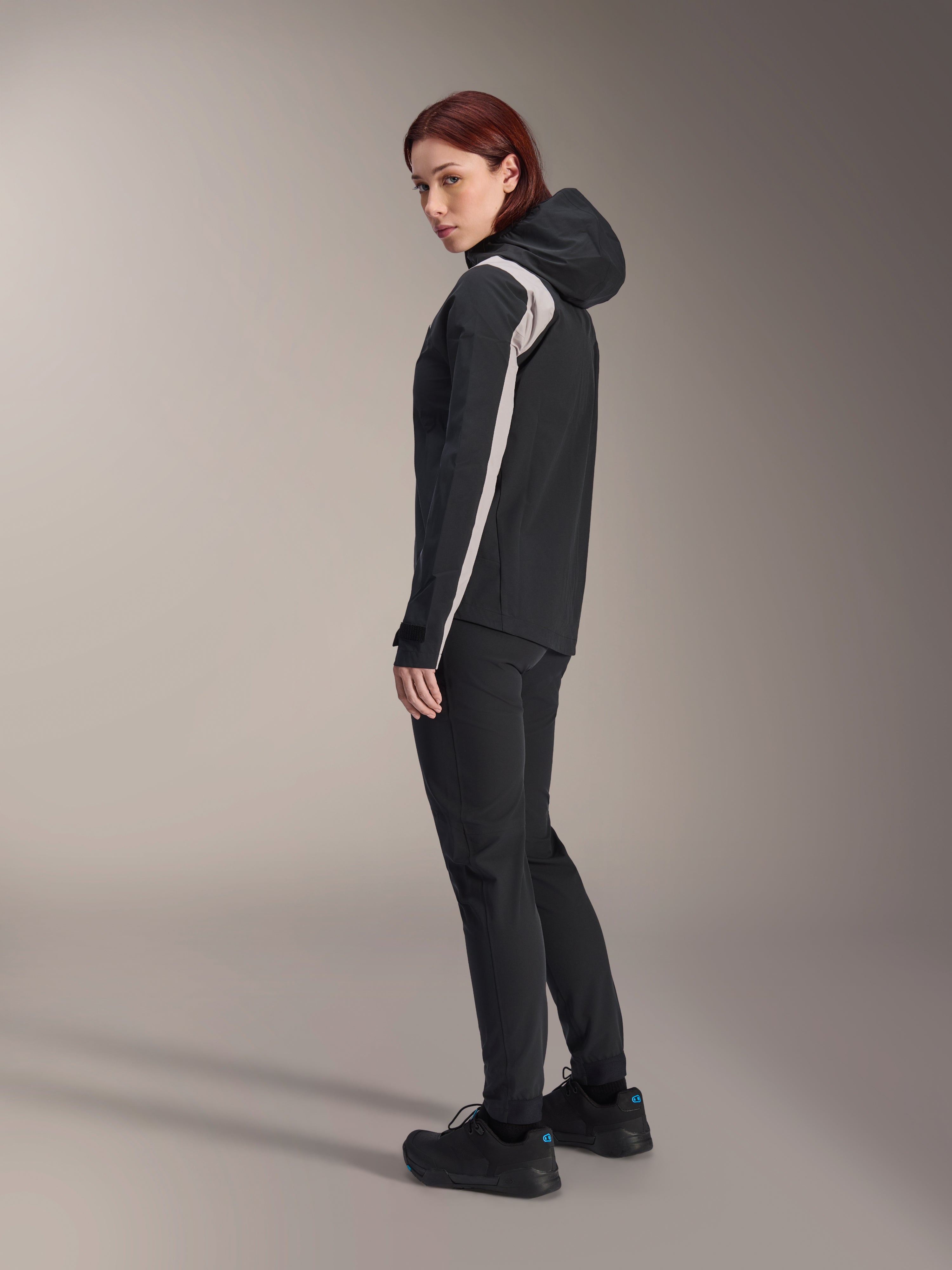 Veste de pluie Alpinestars Stella A-Dura pour femme, noire avec une bande gris clair sur la manche, veste de cyclisme imperméable à capuche pour le VTT, associée à un pantalon technique noir et des chaussures de vélo noires, vue de profil en studio