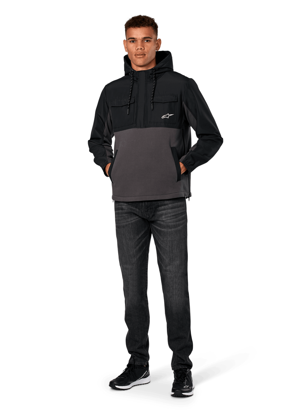 Alpinestars Juncture Hybrid Jas, zwart en gunmetal, anorak hoodie van gemengd materiaal met een romp van canvas fleece en ripstop overlays, wit A-star logo op de borst, capuchon met trekkoord, gedragen door model met donkere jeans en zwarte sneakers, casual motorsport-geïnspireerde streetwear