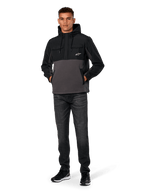 Alpinestars Juncture Hybrid Jas, zwart en gunmetal, anorak hoodie van gemengd materiaal met een romp van canvas fleece en ripstop overlays, wit A-star logo op de borst, capuchon met trekkoord, gedragen door model met donkere jeans en zwarte sneakers, casual motorsport-geïnspireerde streetwear