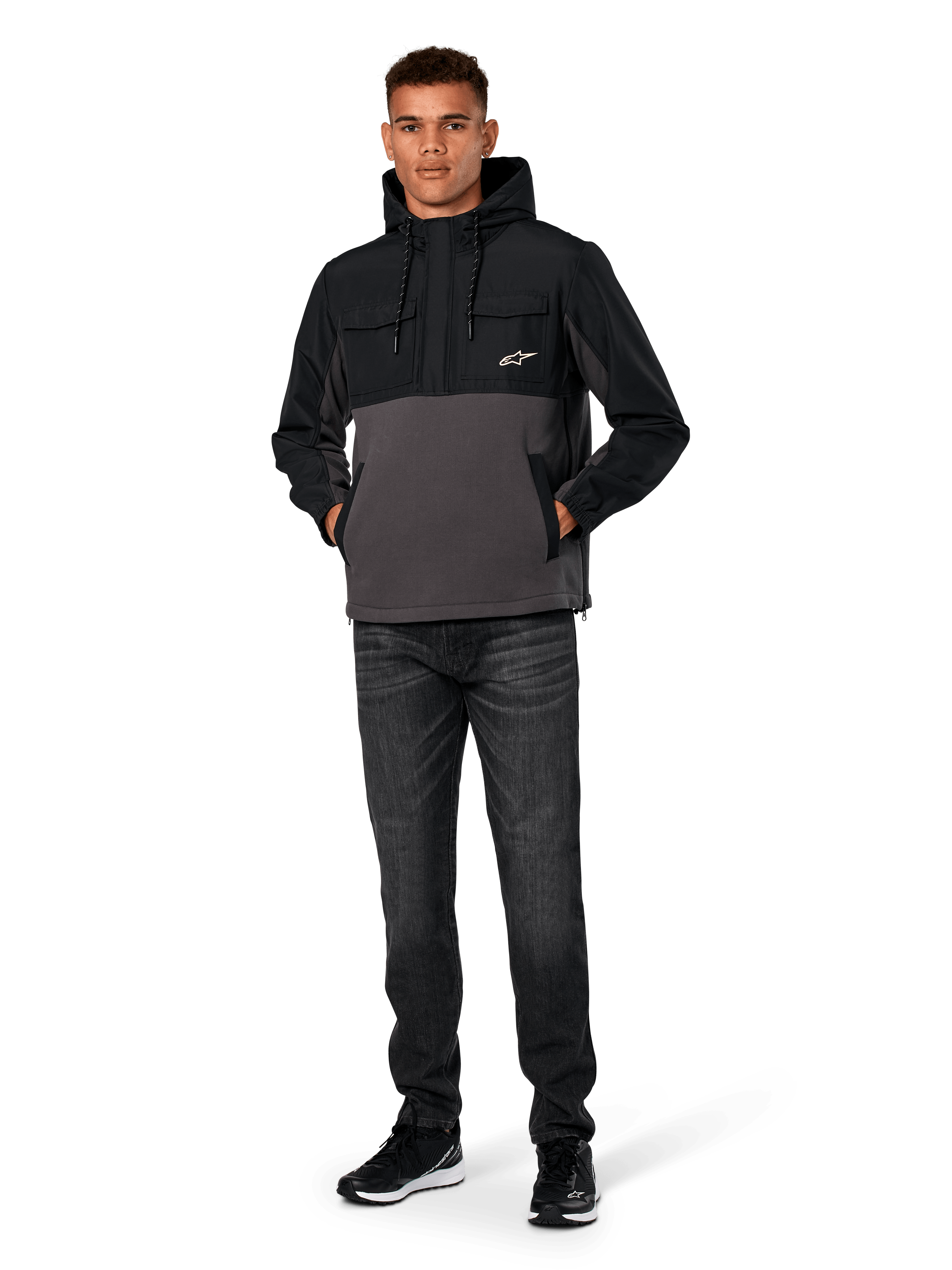 Alpinestars Juncture Hybrid Jas, zwart en gunmetal, anorak hoodie van gemengd materiaal met een romp van canvas fleece en ripstop overlays, wit A-star logo op de borst, capuchon met trekkoord, gedragen door model met donkere jeans en zwarte sneakers, casual motorsport-geïnspireerde streetwear
