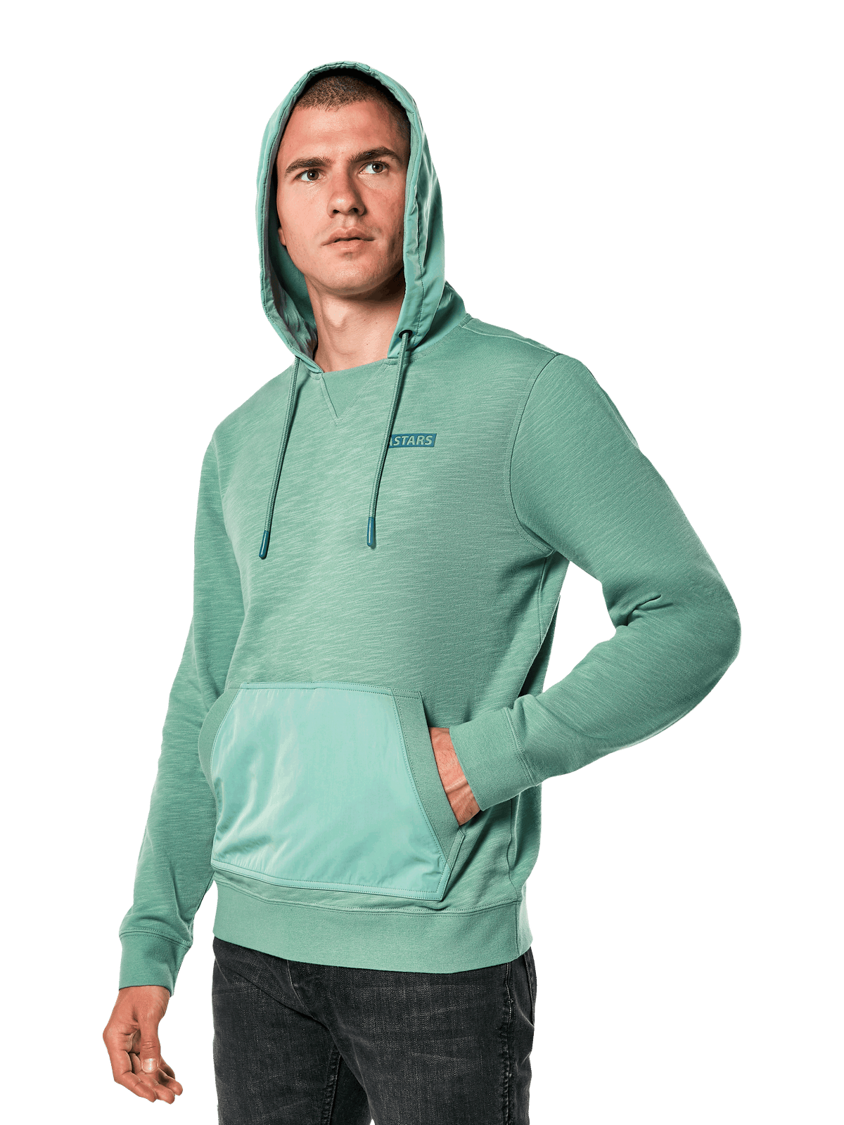 Alpinestars Rooted Hoodie, groene slub French terry hoodie met overlay capuchon en kangoeroezak, klein STARS-logo op de borst, gedragen door een model met de capuchon op en hand in de zak, casual motorsport-geïnspireerde streetwear