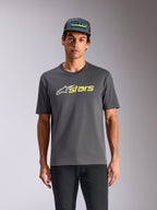 Alpinestars Blaze 2.0 CSF Tee, T-shirt met korte mouwen, antracietgrijs, wit ster-logo en felgele stars-graphic, gedragen met bijpassende truckerpet en zwarte broek, casual motorsport streetwear