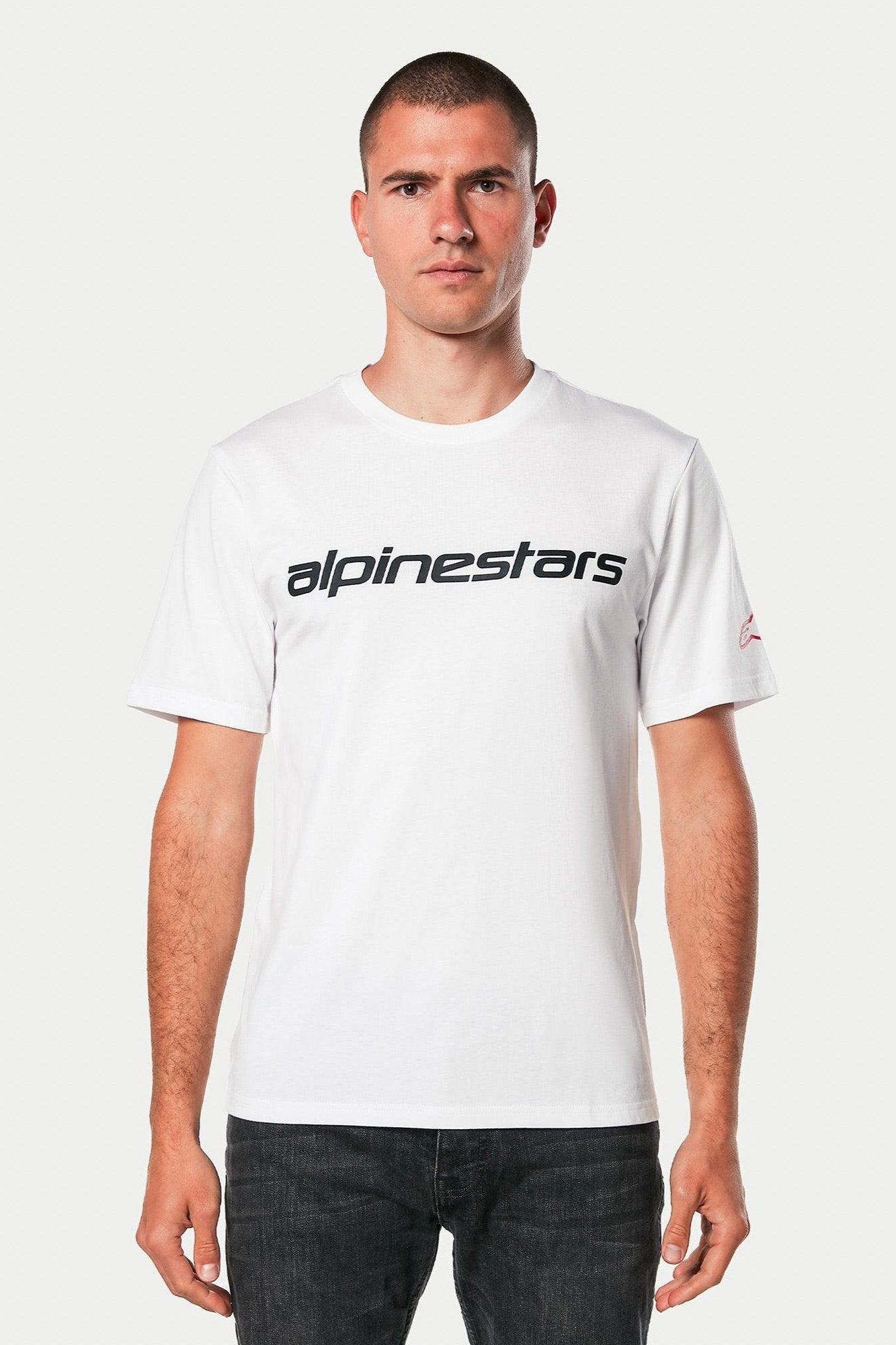 Alpinestars Linear Wordmark 2.0 CSF Tee, Kurzarm-T-Shirt, Weiß mit schwarzem linearem Schriftzug auf der Brust und rotem Logo am Ärmel, von einem Model mit dunklen Jeans getragen, lässige Motorsport-Streetwear