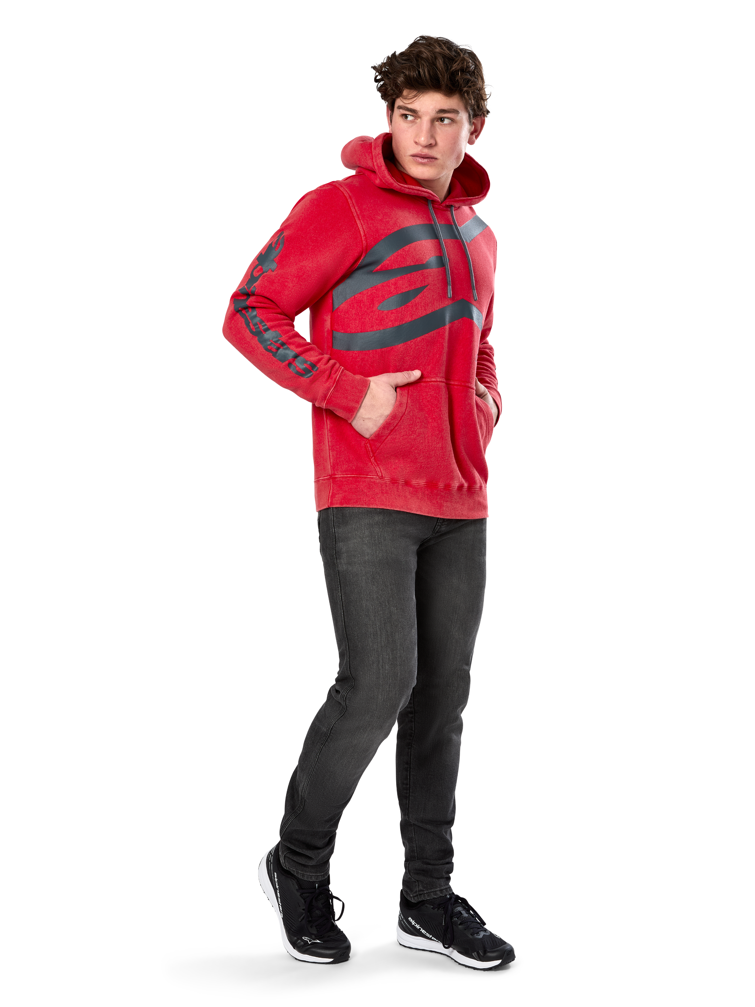 Alpinestars Unbound Hoodie, rode kledinggeverfde pullover fleece, extra groot donker logo op de borst en woordmerk op de mouwen, kangoeroezak, capuchon met trekkoord, gedragen door model met donkergrijze jeans en zwarte sneakers, casual op motorsport geïnspireerde streetwear