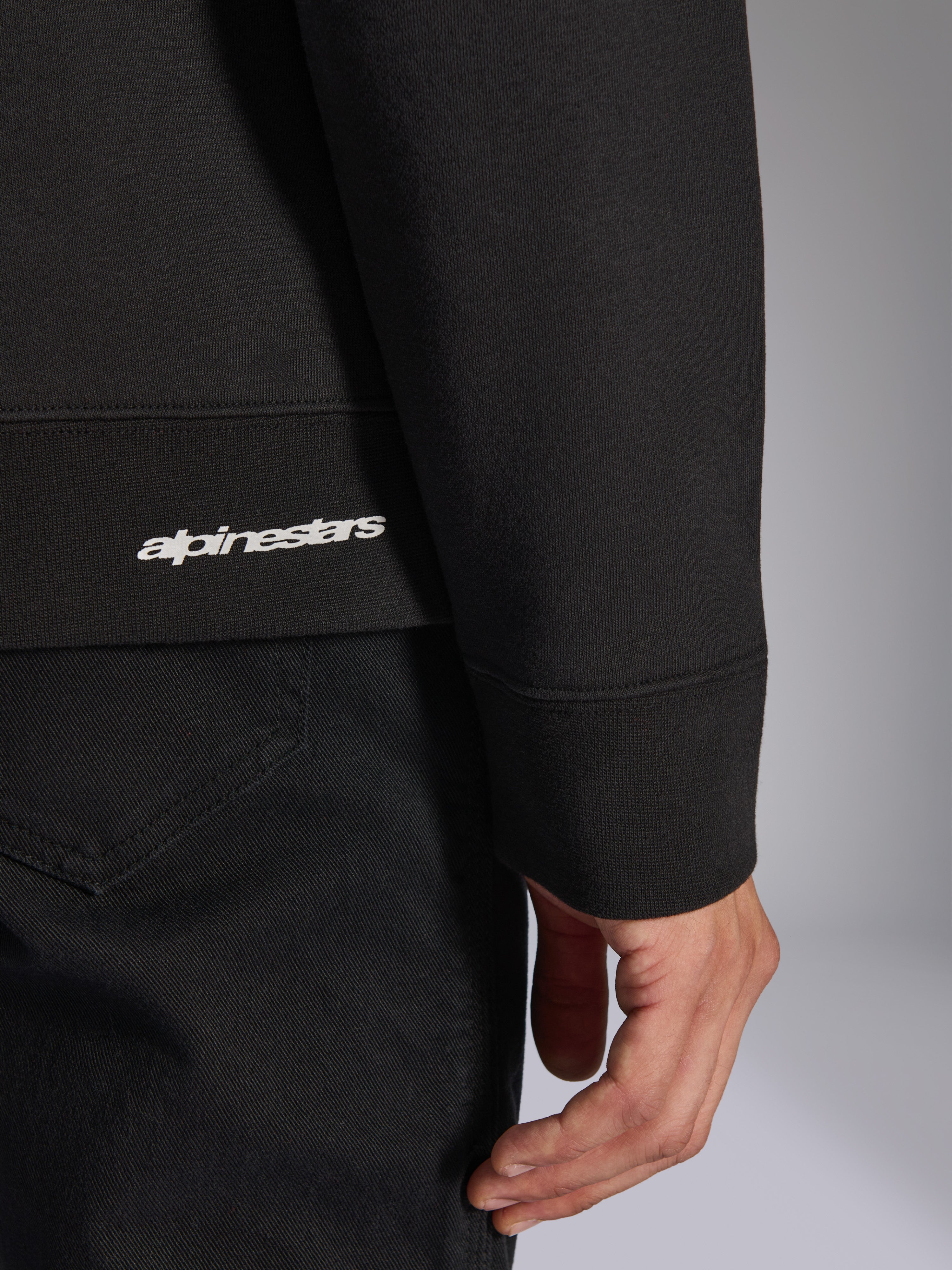 Alpinestars Positive Crew, sweatshirt noir, gros plan sur l'ourlet côtelé et le poignet avec logo Alpinestars blanc, texture de tissu premium, vêtement de sport décontracté d'inspiration sport automobile