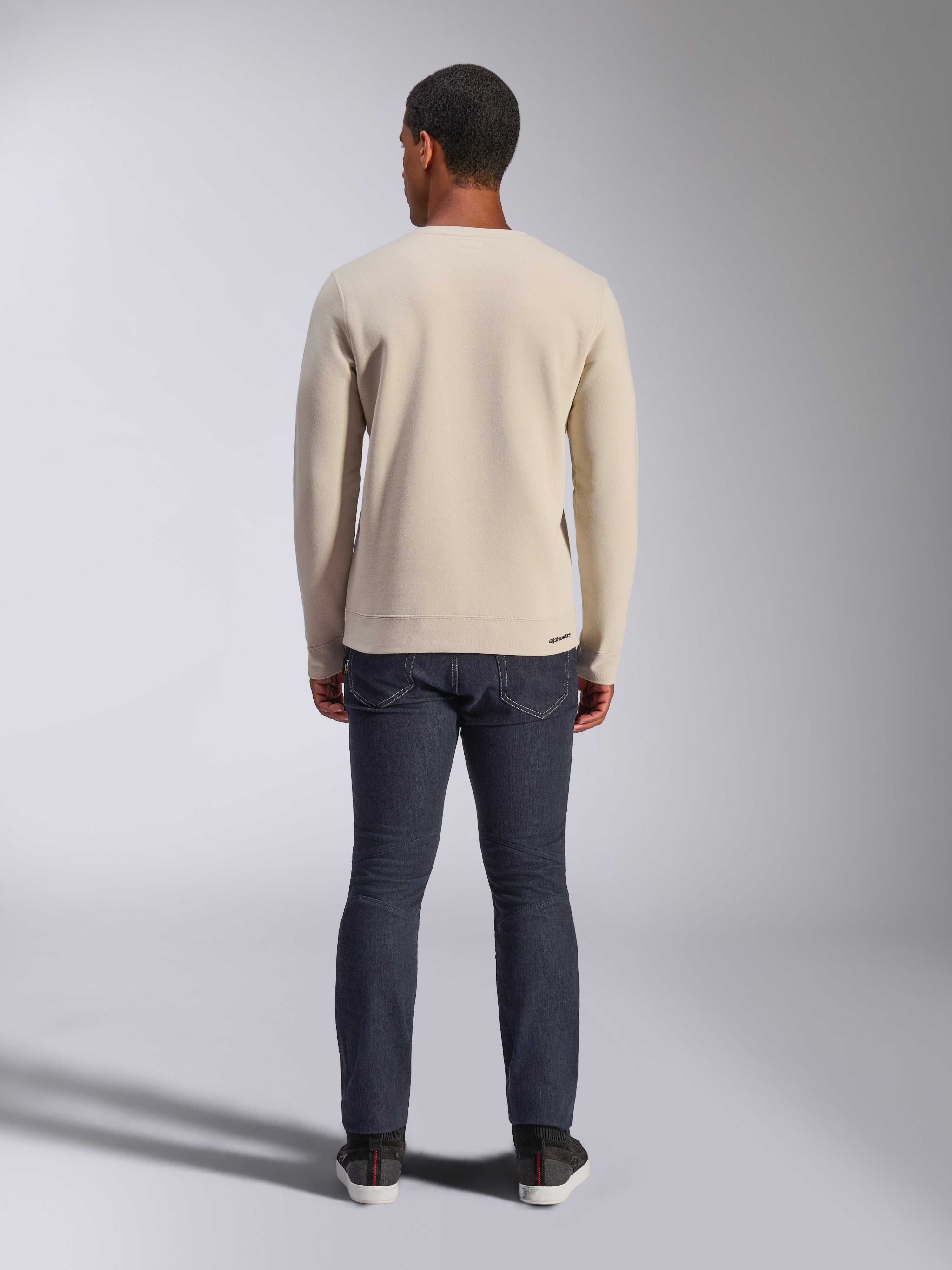 Alpinestars Positive Crew Sweatshirt, Off White, achteraanzicht van een man die een beige pullover met ronde hals en geribbelde zoom en manchetten draagt, gecombineerd met een donkere jeans, casual motorsport-geïnspireerde streetwear.