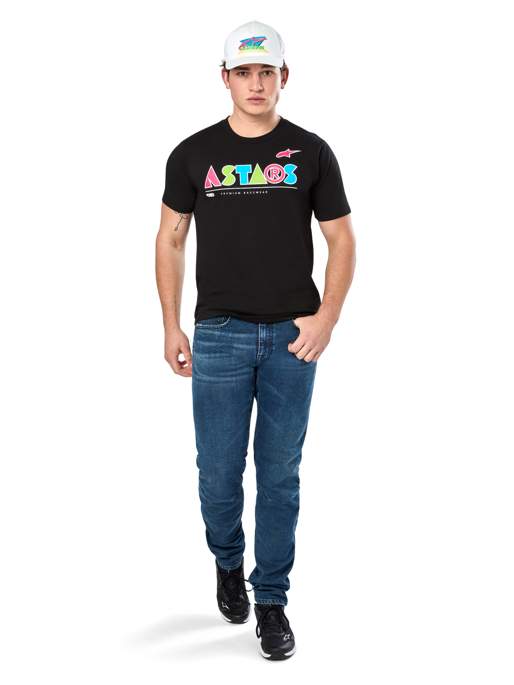 Mannelijk model draagt Alpinestars Cereal CSF Tee, zwart T-shirt met korte mouwen met kleurrijk geometrisch ASTARS-logo, gecombineerd met blauwe jeans en witte pet, casual motorsport-geïnspireerde streetwear.