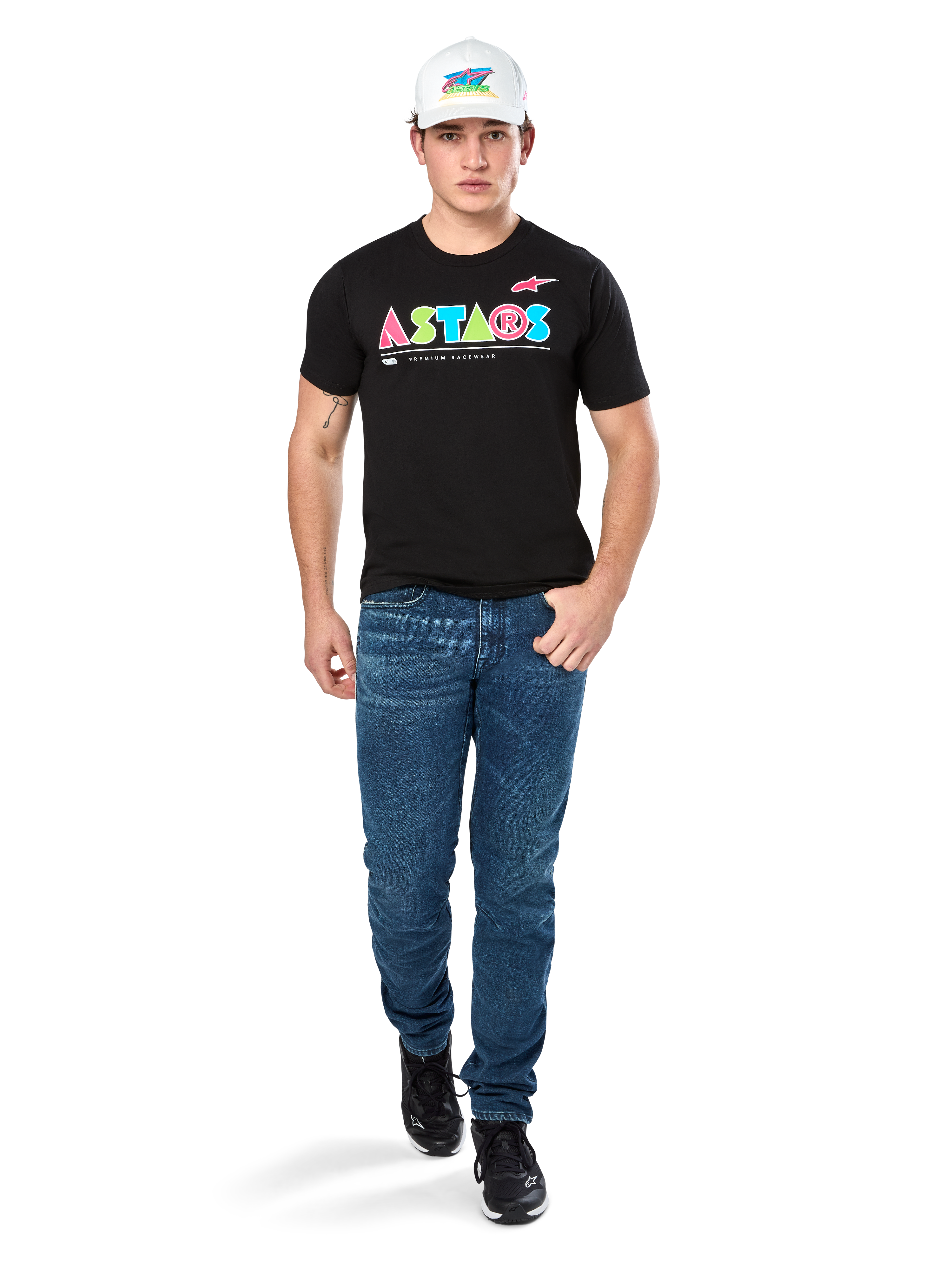Mannelijk model draagt Alpinestars Cereal CSF Tee, zwart T-shirt met korte mouwen met kleurrijk geometrisch ASTARS-logo, gecombineerd met blauwe jeans en witte pet, casual motorsport-geïnspireerde streetwear.