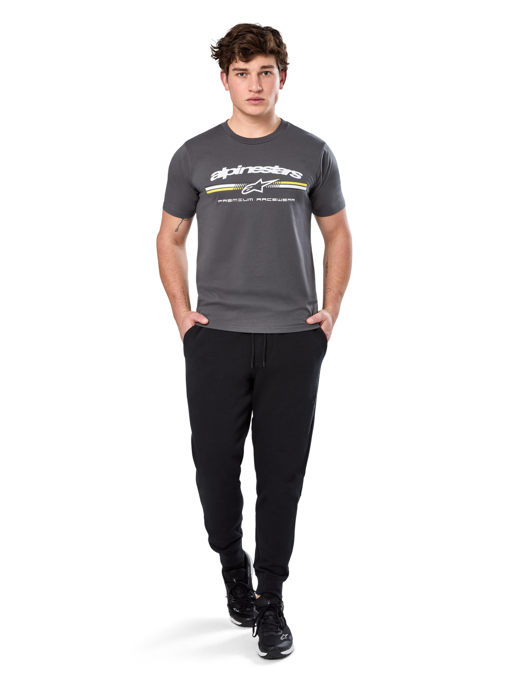 Man in Alpinestars Prevail Csf Tee, T-shirt met korte mouwen, charcoal, voorzien van wit Alpinestars-logo en driekleurige streep met gele accenten, gecombineerd met zwarte joggingbroek en sneakers, casual motorsport streetwear