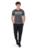 Man in Alpinestars Prevail Csf Tee, T-shirt met korte mouwen, charcoal, voorzien van wit Alpinestars-logo en driekleurige streep met gele accenten, gecombineerd met zwarte joggingbroek en sneakers, casual motorsport streetwear