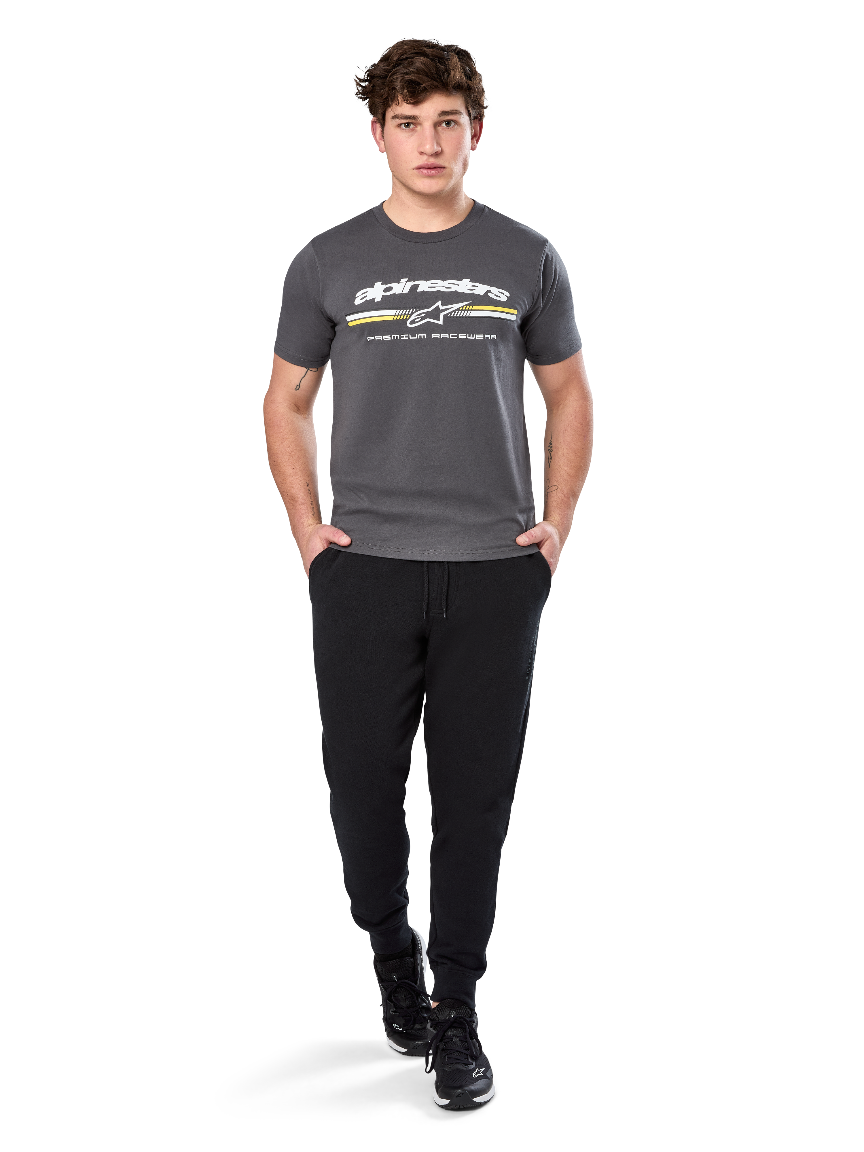 Man in Alpinestars Prevail Csf Tee, T-shirt met korte mouwen, charcoal, voorzien van wit Alpinestars-logo en driekleurige streep met gele accenten, gecombineerd met zwarte joggingbroek en sneakers, casual motorsport streetwear