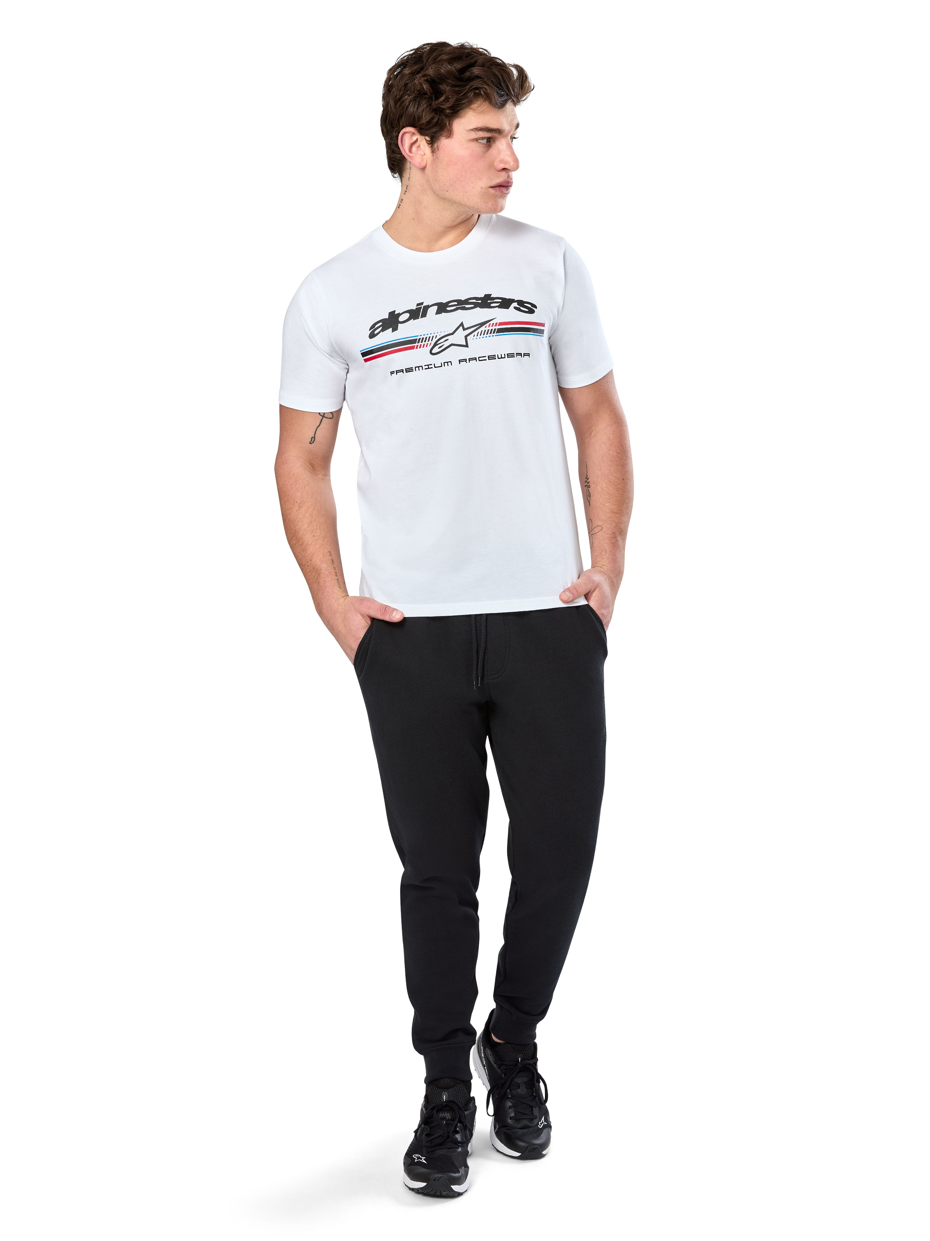 Alpinestars Prevail CSF Tee, T-shirt met korte mouwen, wit, met een driekleurig logo in paddock-stijl op de borst en Alpinestars-branding, gedragen door een model met een zwarte joggingbroek en zwarte sneakers, casual motorsport streetwear