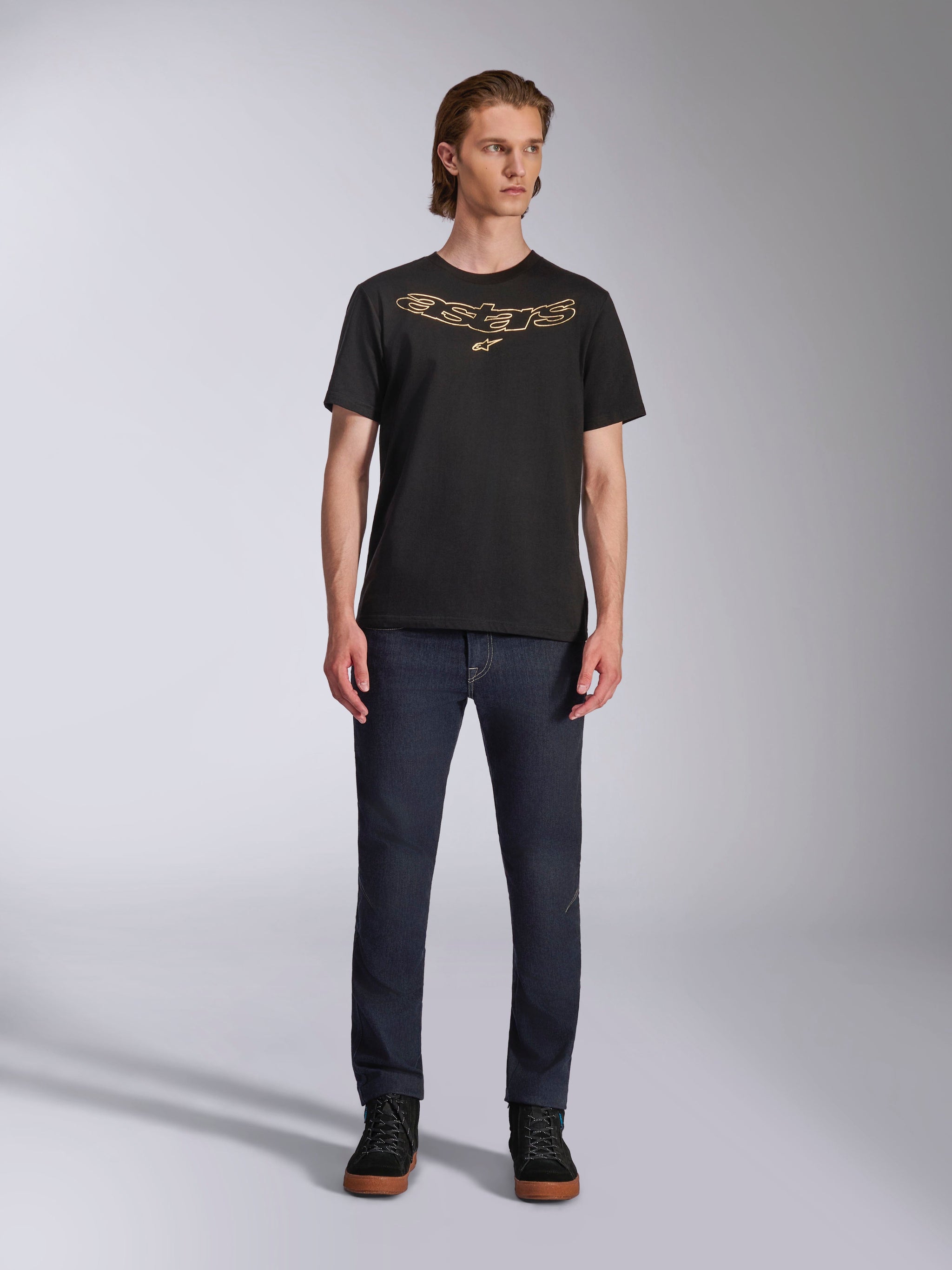 Alpinestars Luxo CSF Tee, T-shirt met korte mouwen, zwart, Astars-logo met beige omlijning op de borst, gedragen door model met donkerblauwe denim jeans en zwarte hoge schoenen, casual motorsport streetwear