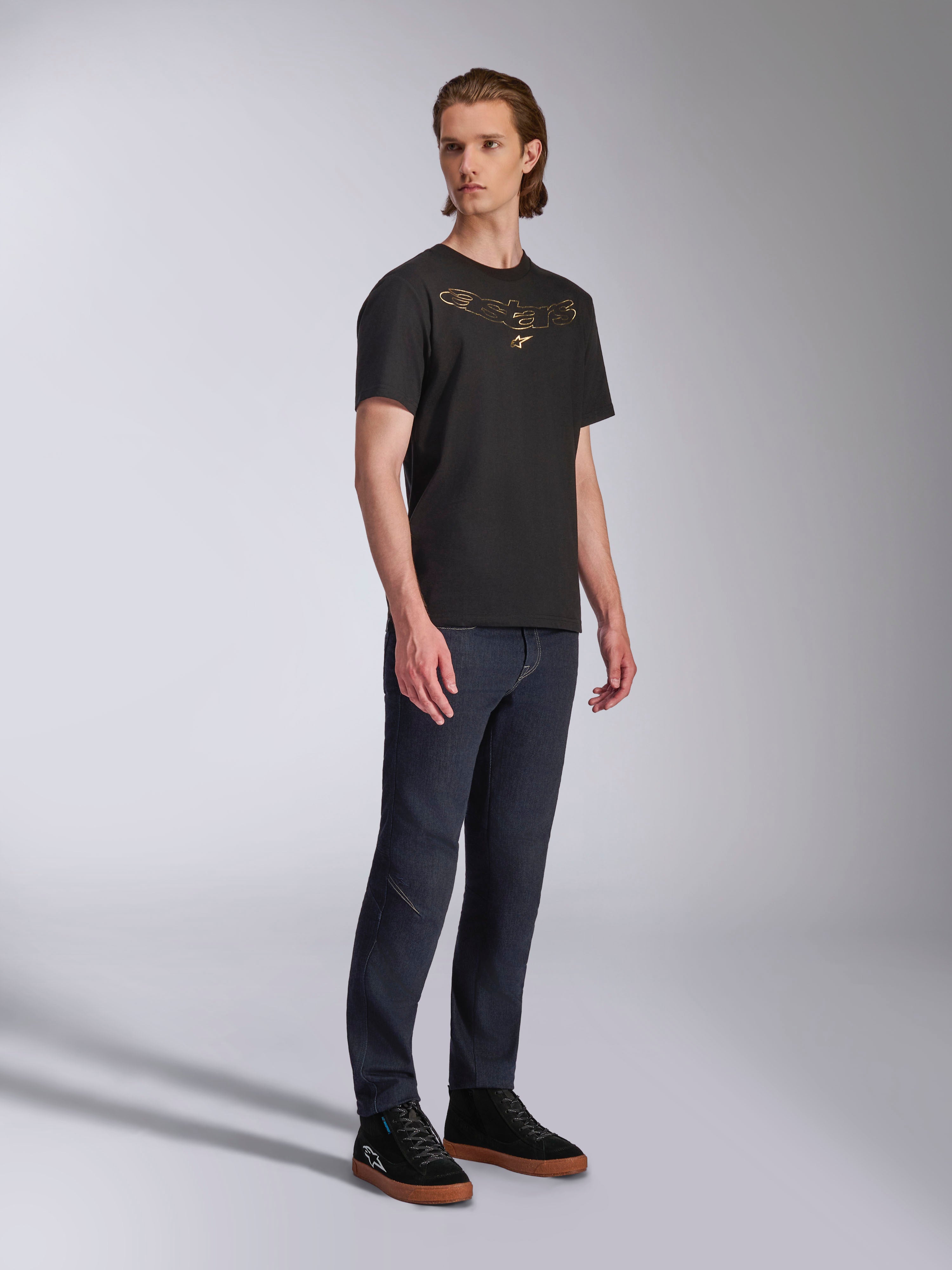 Alpinestars Luxo CSF T-shirt, T-shirt met korte mouwen, zwart, goudkleurig omlijnd Astars-logo op de borst, gedragen door model met donkere denim jeans en zwarte sneakers, casual motorsport streetwear