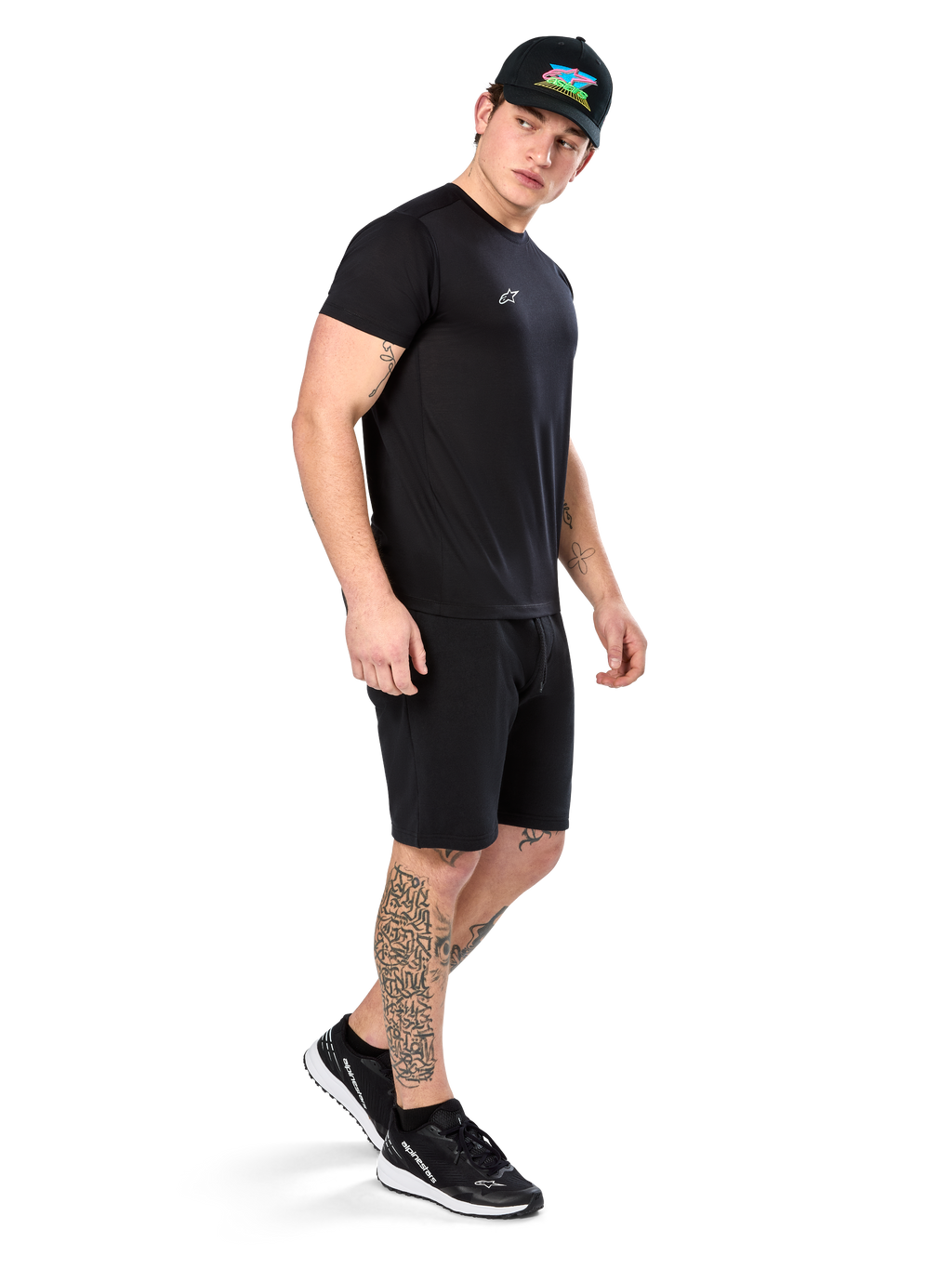 Man met tatoeages in Alpinestars Point Performance Tee, zwart performance T-shirt met korte mouwen, klein wit logo, gecombineerd met zwarte shorts, een zwarte pet met kleurrijke graphic en zwart-witte sneakers, sportieve casual lifestyle
