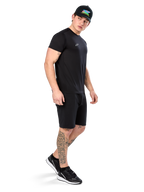 Man met tatoeages in Alpinestars Point Performance Tee, zwart performance T-shirt met korte mouwen, klein wit logo, gecombineerd met zwarte shorts, een zwarte pet met kleurrijke graphic en zwart-witte sneakers, sportieve casual lifestyle