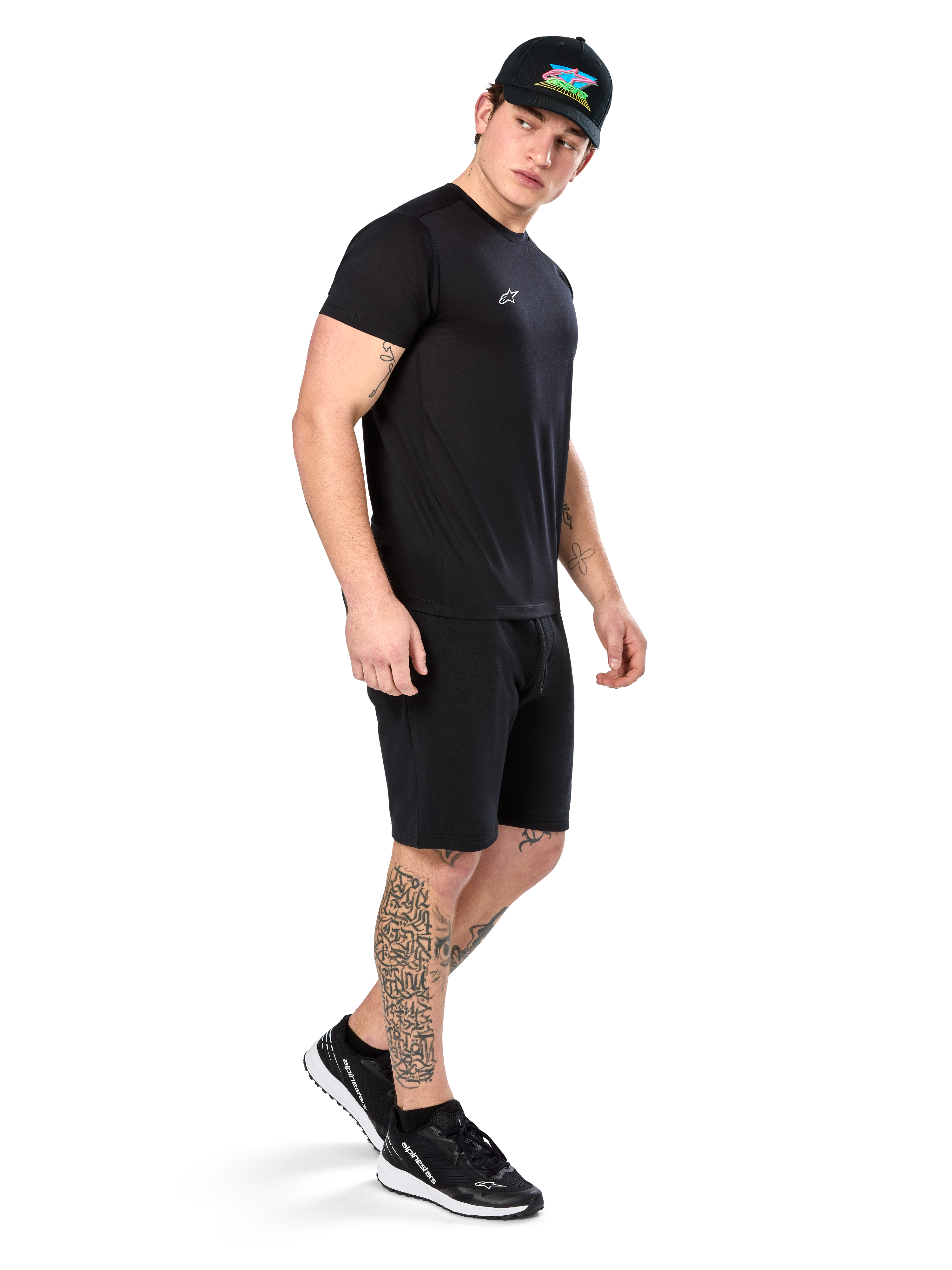 Man met tatoeages in Alpinestars Point Performance Tee, zwart performance T-shirt met korte mouwen, klein wit logo, gecombineerd met zwarte shorts, een zwarte pet met kleurrijke graphic en zwart-witte sneakers, sportieve casual lifestyle