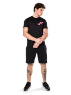Model draagt Alpinestars Attrition Performance Tee, performance T-shirt met korte mouwen, zwart met een roze Astars-logo op de borst, gecombineerd met zwarte shorts en sneakers, casual op motorsport geïnspireerde sportkleding