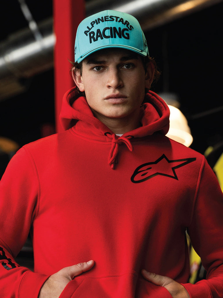 Alpinestars Luminary Kappe, Snapback in Aqua-Blau mit gebogenem Schirm, schwarze 3D-Stickerei mit „ALPINESTARS RACING“-Schriftzug, getragen mit einem leuchtend roten Alpinestars-Hoodie, lässige Motorsport-Lifestyle-Streetwear