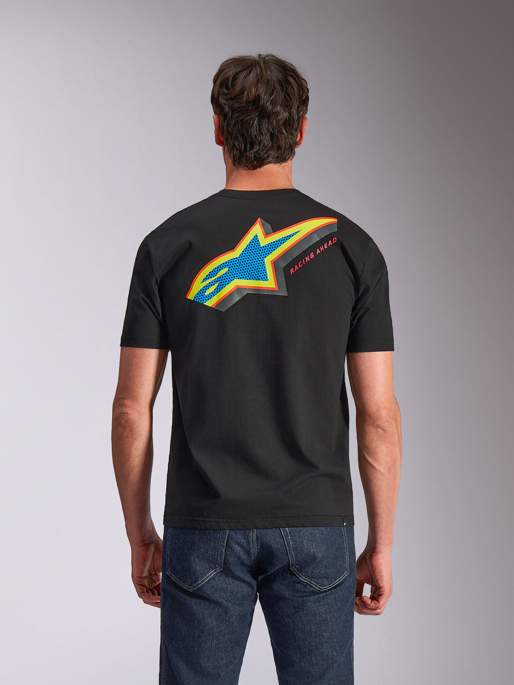 Alpinestars Angler CSF Tee, T-shirt met korte mouwen, zwart, achteraanzicht met levendig meerkleurig Astars-logo met geperforeerde print en de tekst 'RACING AHEAD', gedragen met een blauwe jeans, casual motorsport-geïnspireerde kleding