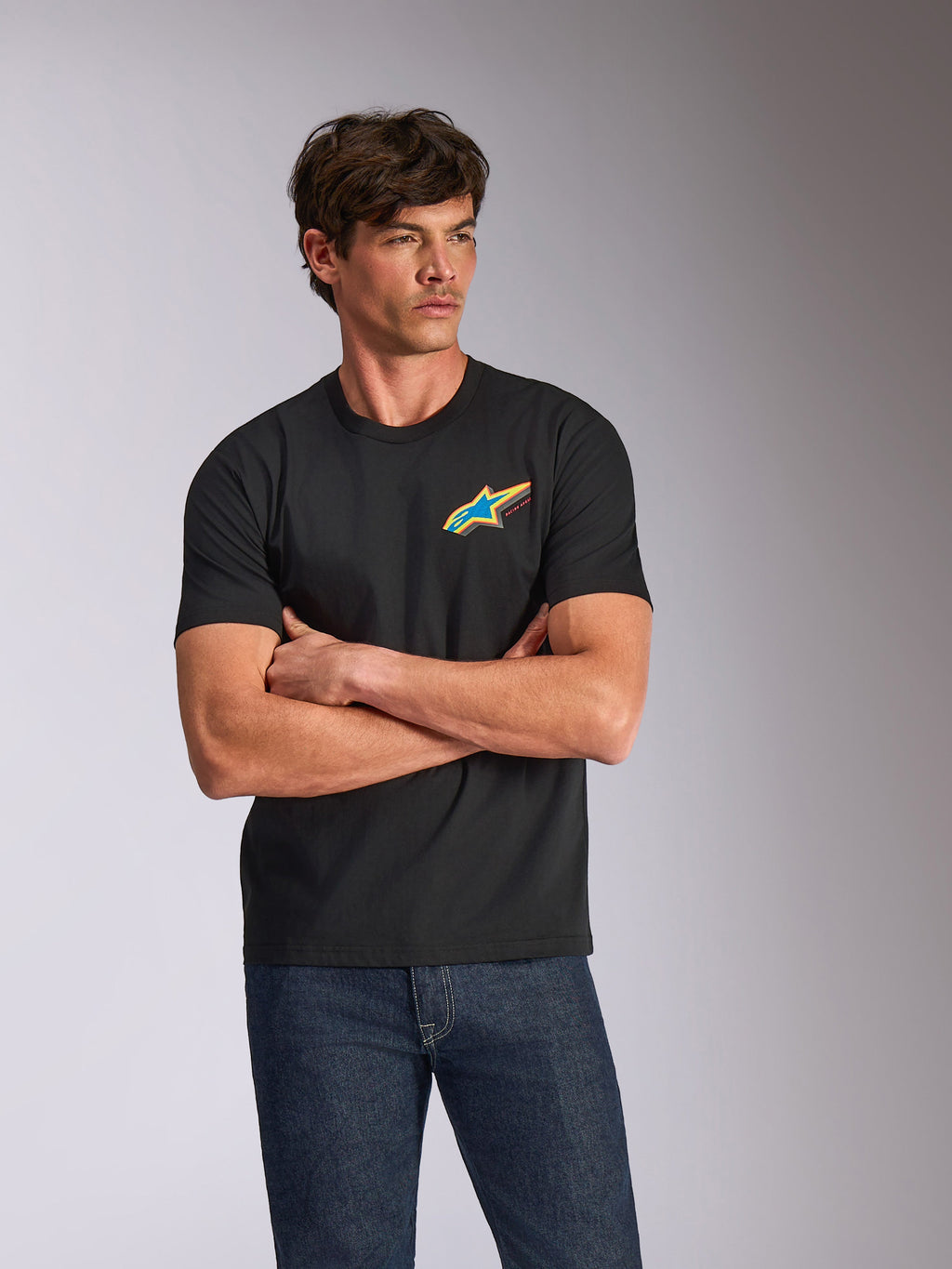 Alpinestars Angler CSF Tee, T-shirt met korte mouwen, zwart, voorzien van een levendig meerkleurig Astars-logo op de borst, gedragen door een model met de armen over elkaar en een blauwe spijkerbroek, casual motorsport-geïnspireerde streetwear