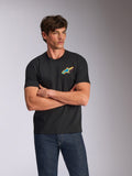 Alpinestars Angler CSF Tee, T-shirt met korte mouwen, zwart, voorzien van een levendig meerkleurig Astars-logo op de borst, gedragen door een model met de armen over elkaar en een blauwe spijkerbroek, casual motorsport-geïnspireerde streetwear