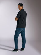 Alpinestars Impetus Sport Polo, zwart, poloshirt met korte mouwen met mock-mesh textuur en knoopsluiting met drie knopen, gedragen door een model met blauwe jeans en zwarte sneakers, casual streetwear geïnspireerd op de motorsport