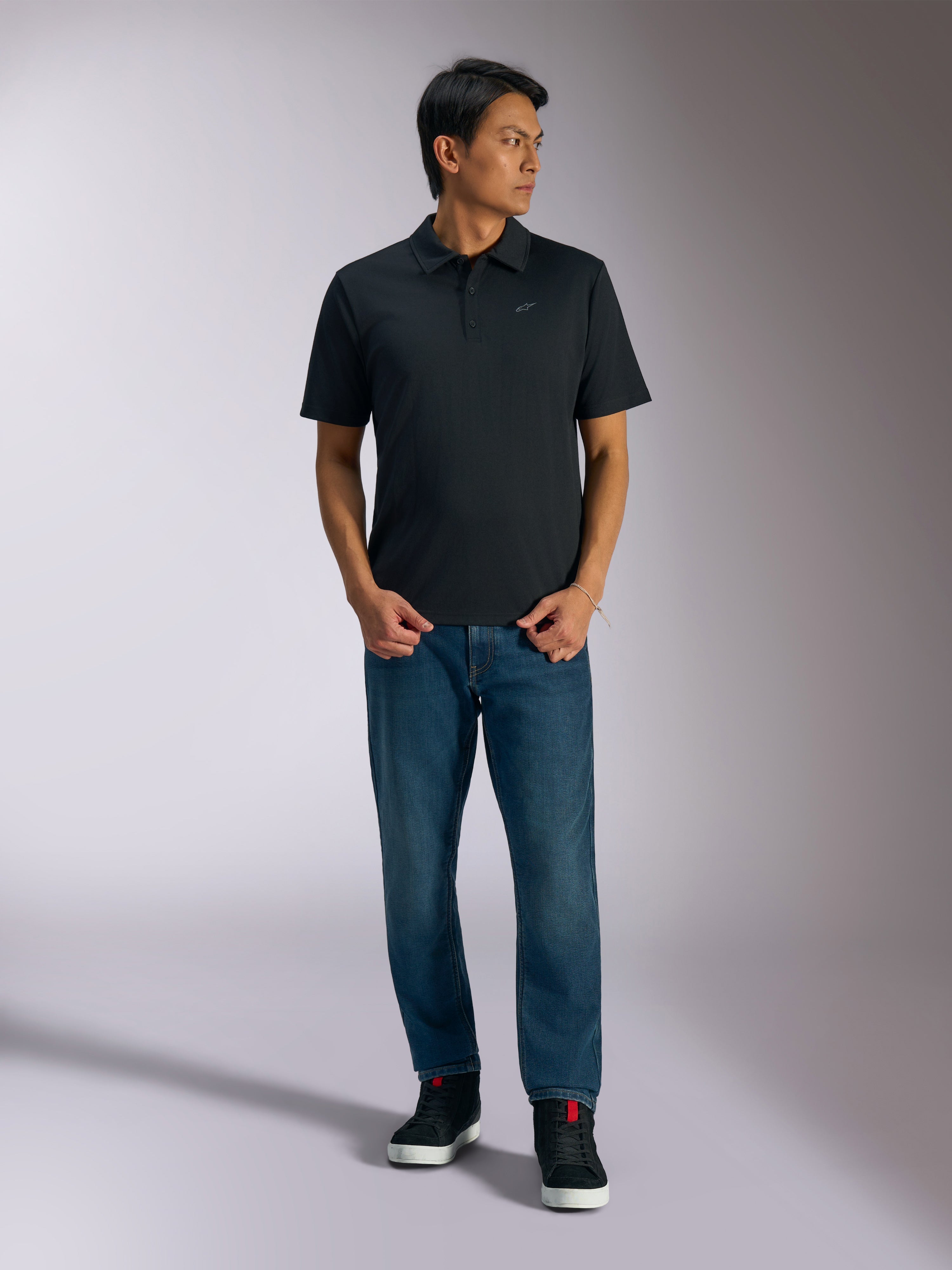 Alpinestars Impetus Sport Polo, zwart poloshirt met korte mouwen, voorzien van een knoopsluiting met drie knopen en een grijs Astars-logo op de borst, gedragen door een man met een blauwe jeans en zwarte hoge sneakers, casual motorsport-geïnspireerde sportkleding