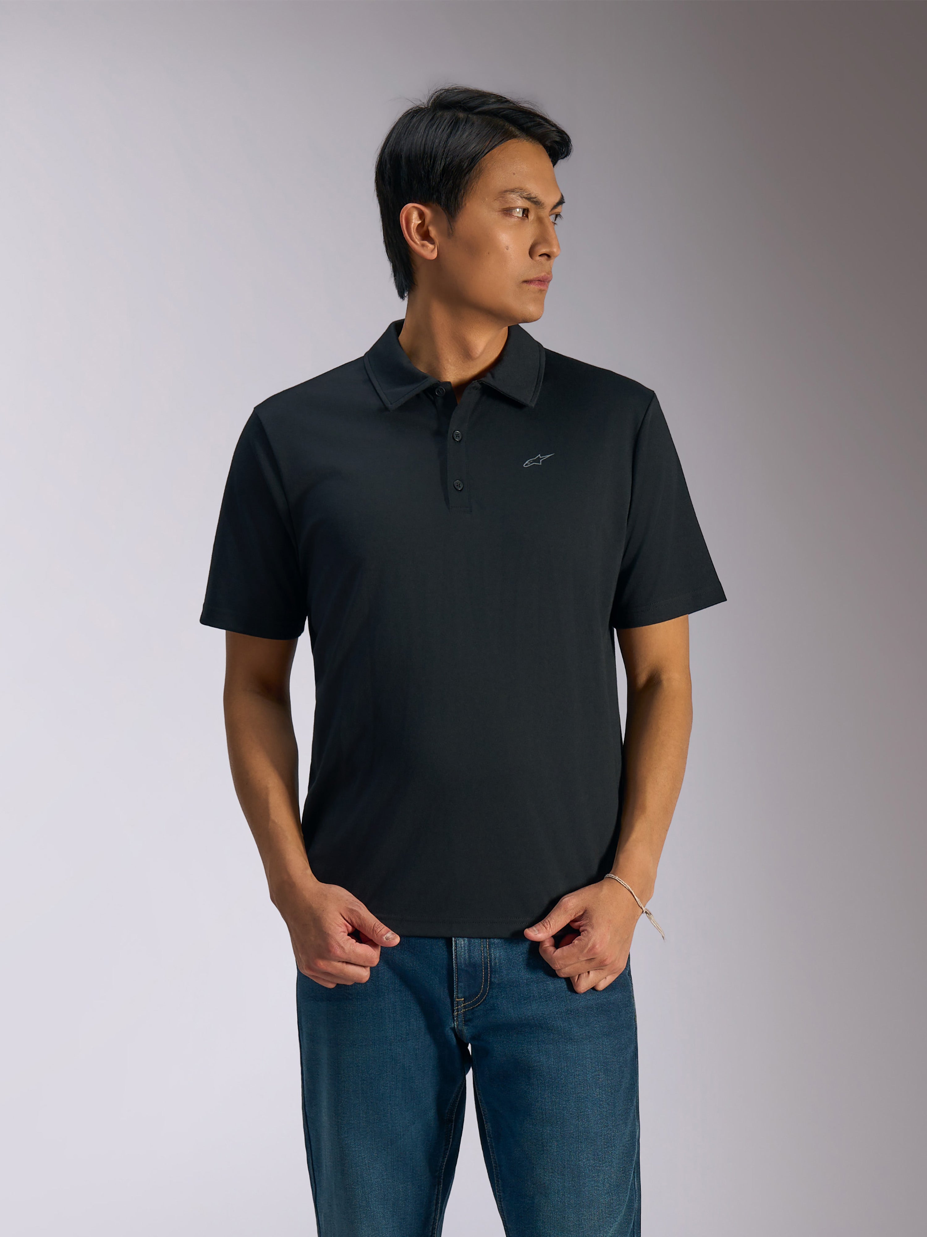Alpinestars Impetus Sport Polo, zwarte polo met korte mouwen, sluiting met drie knopen en zelfkraag, klein grijs Astars-logo op de linkerborst, gedragen met een blauwe jeans, casual motorsport-geïnspireerde sportkleding