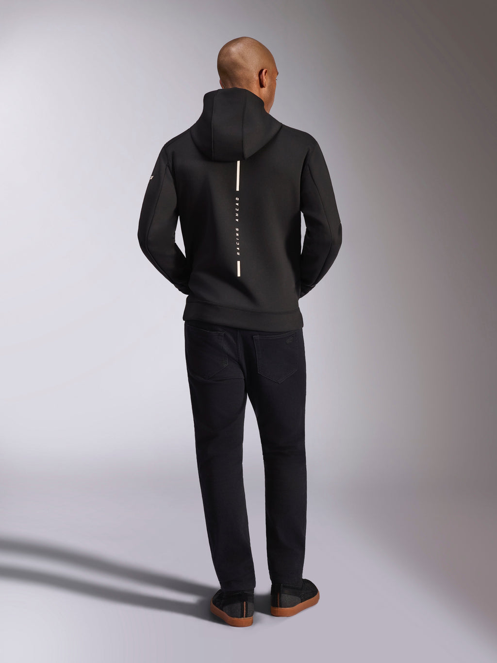 Alpinestars Insight Hoodie, zwart, achteraanzicht op model, voorzien van een verticaal wit streep-logo op de rug met de tekst "Racing Ahead", casual sportswear hoodie van tri-blend double-face fleece, gecombineerd met een zwarte broek.