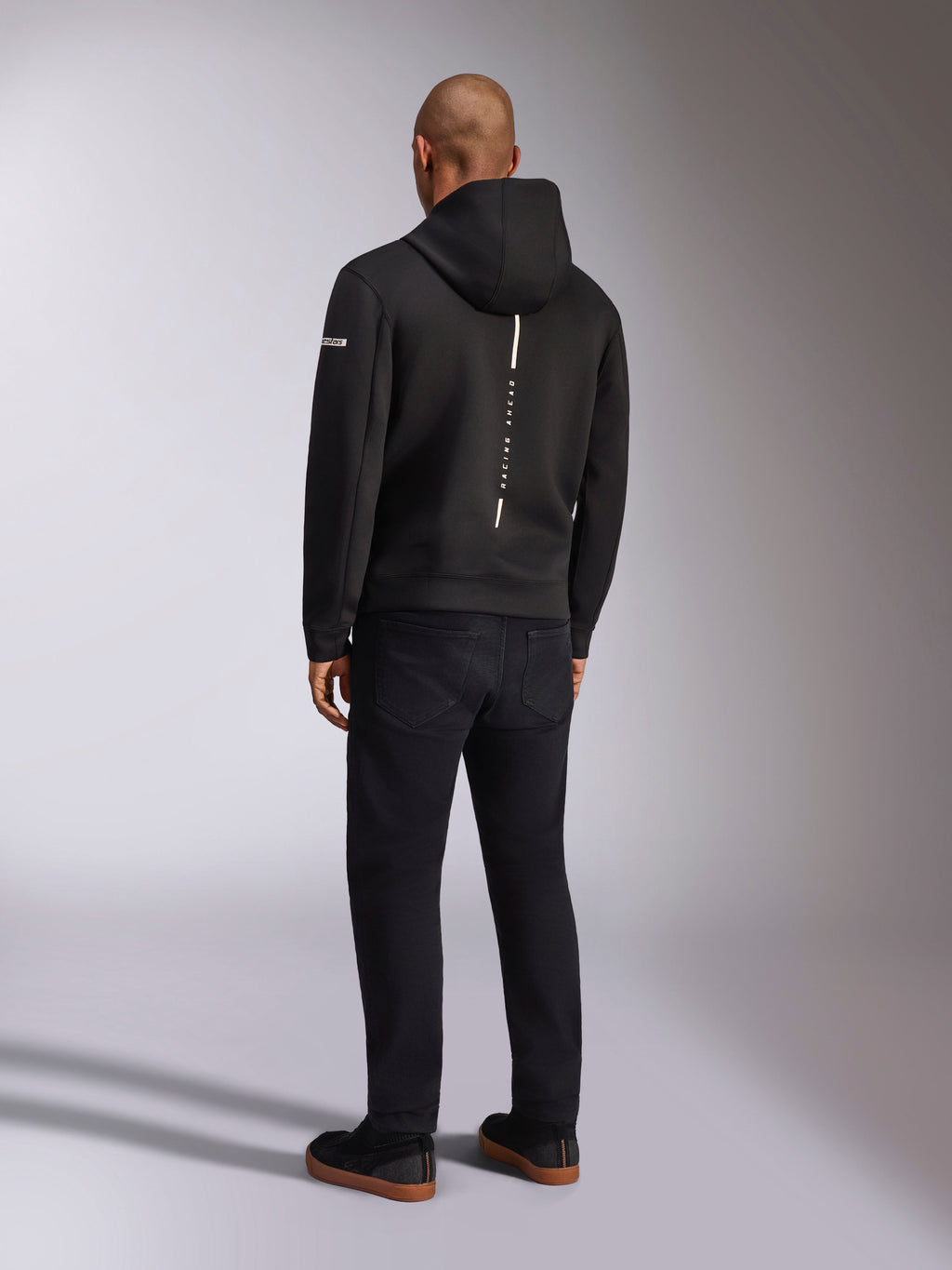 Alpinestars Insight Hoodie, zwarte sportswear hoodie, achteraanzicht met een verticale witte streep en 'RACING AHEAD' branding, logo op de mouw, gecombineerd met een zwarte broek en sneakers, casual op de motorsport geïnspireerd ontwerp