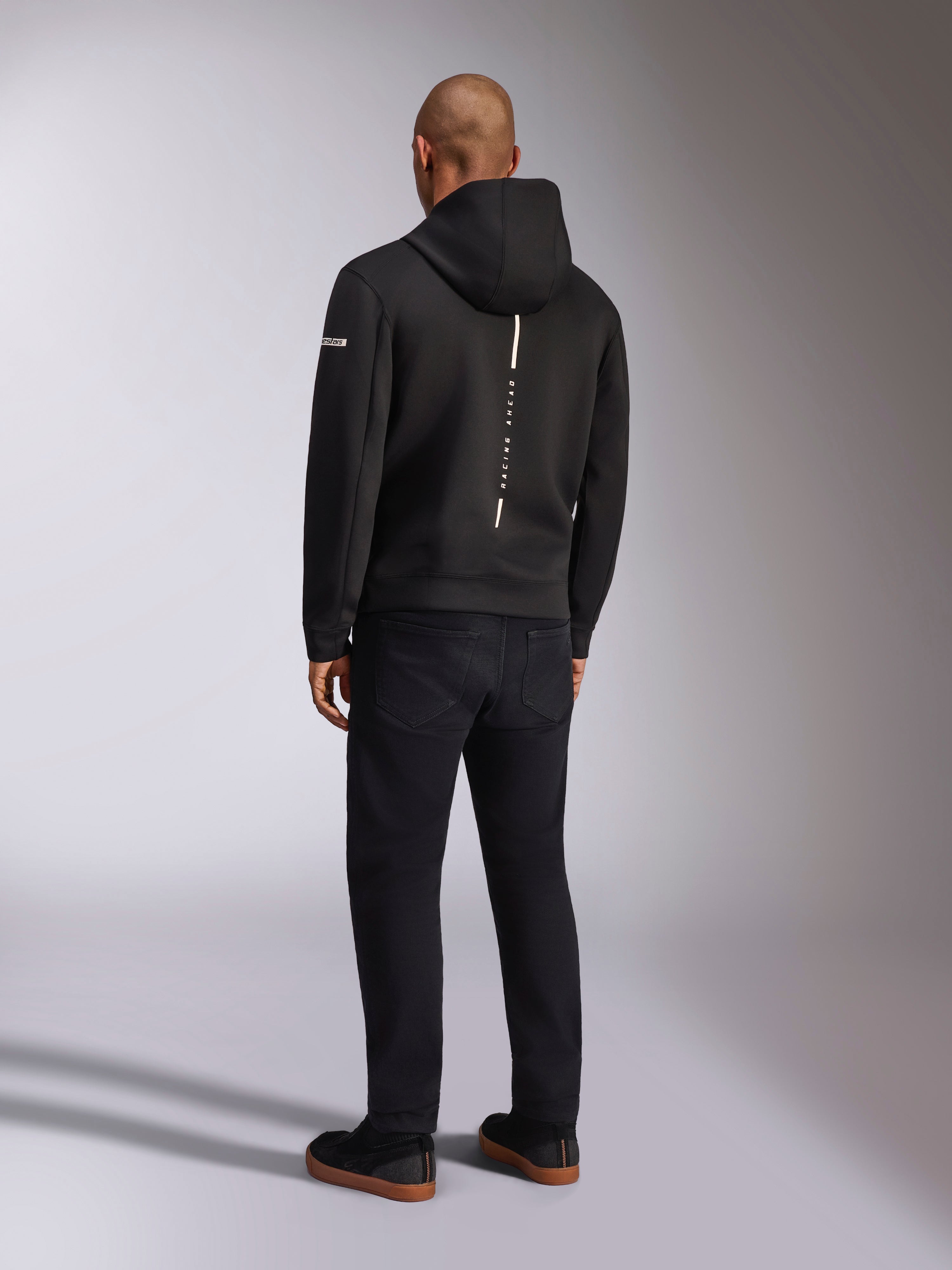 Alpinestars Insight Hoodie, zwarte sportswear hoodie, achteraanzicht met een verticale witte streep en 'RACING AHEAD' branding, logo op de mouw, gecombineerd met een zwarte broek en sneakers, casual op de motorsport geïnspireerd ontwerp
