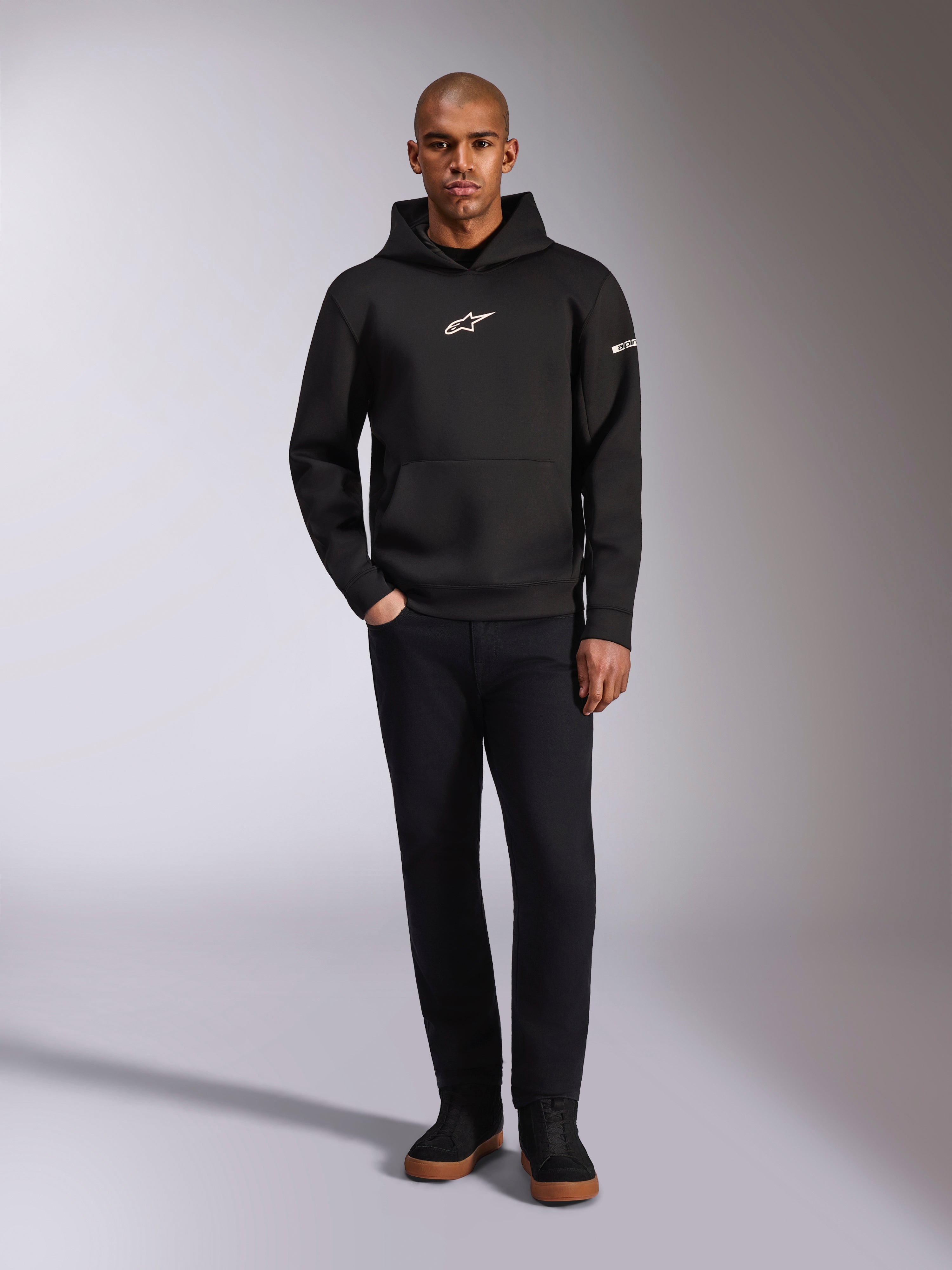 Alpinestars Insight Hoodie, zwarte pullover hoodie met wit Astars-logo op de borst en mouw, gedragen door een model met zwarte broek en zwarte sneakers, casual motorsport-geïnspireerde streetwear
