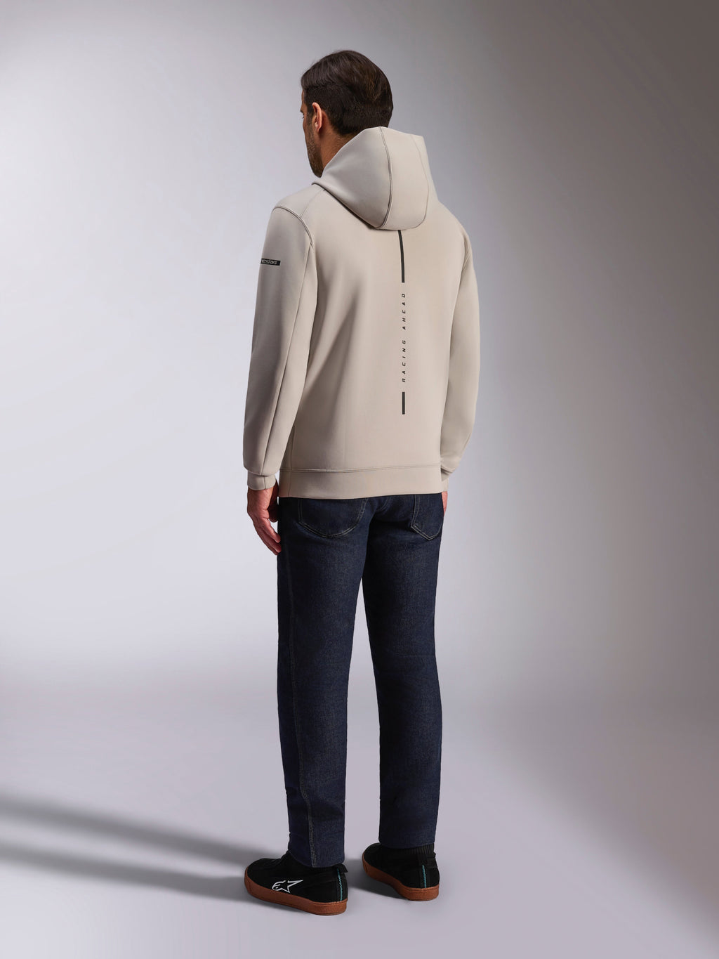 Alpinestars Insight Hoodie, achteraanzicht in cementgrijs, voorzien van een verticale zwarte streep met de tekst 'RACING AHEAD', casual streetwear-ontwerp, gedragen met een donkere jeans en zwarte schoenen.