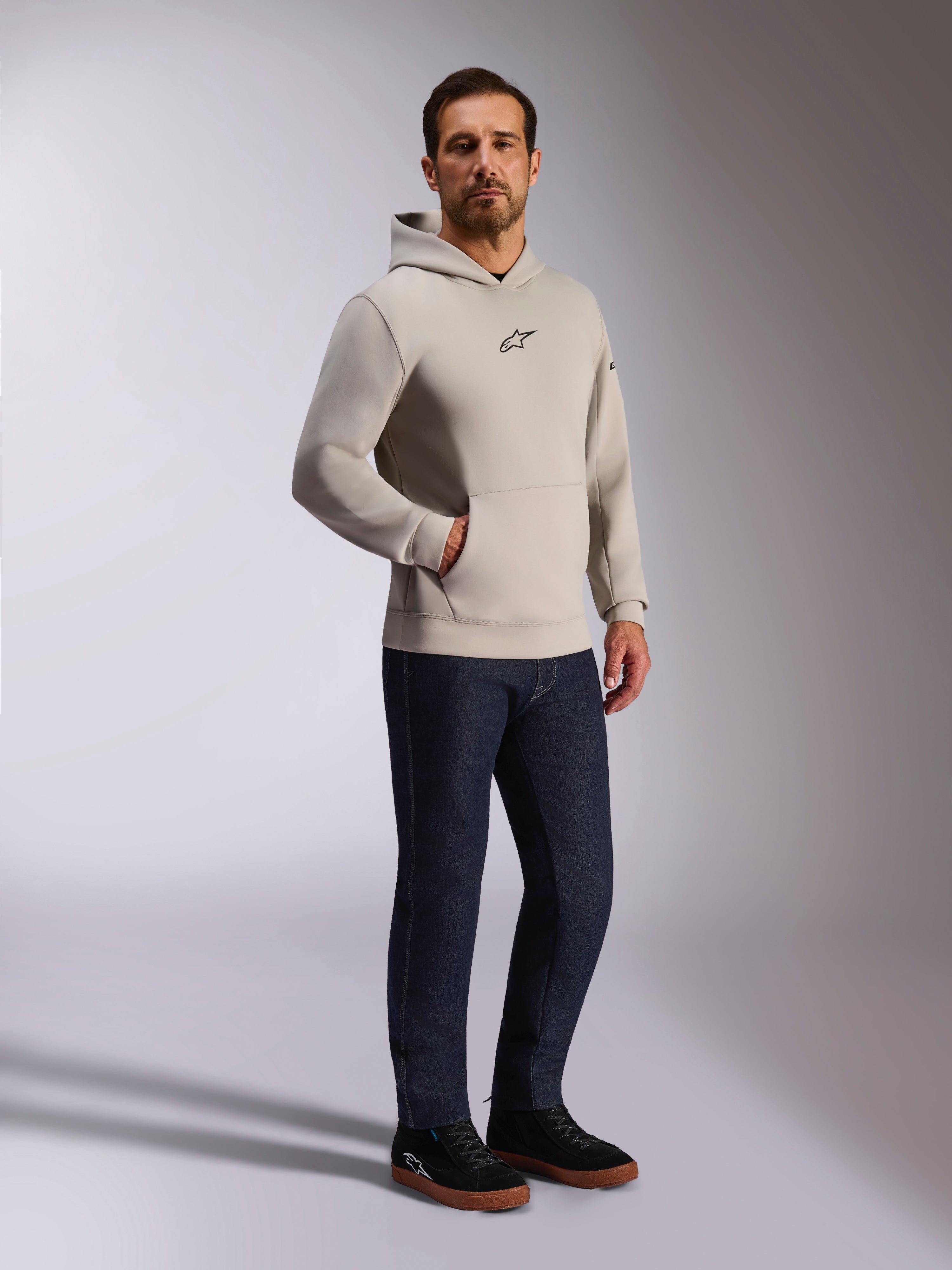 Alpinestars Insight Hoodie, casual streetwear, cementgrijs met zwart Astars-logo op de borst, pullover-ontwerp met kangoeroezak, gedragen met donkerblauwe jeans en zwarte sneakers, moderne op motorsport geïnspireerde stijl
