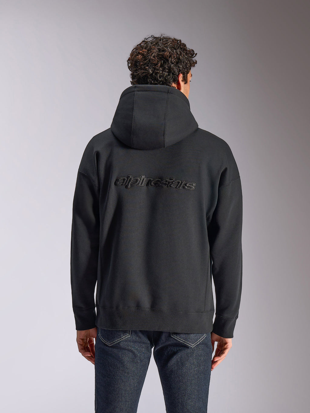 Alpinestars Emergent Oversized Hoodie, achteraanzicht van een zwarte pullover hoodie op een model, met geborduurd Alpinestars-woordlogo op de rug, oversized pasvorm, casual streetwear gecombineerd met een donkere jeans