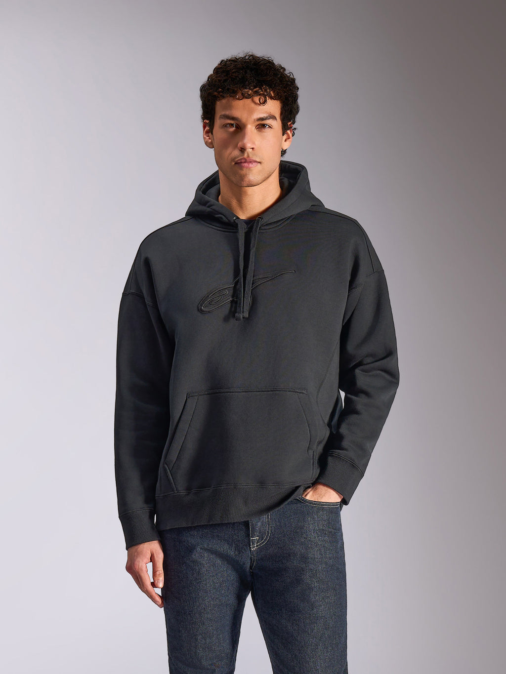Alpinestars Emergent Oversized Hoodie, zwart, mannelijk model draagt een oversized pullover fleece hoodie, tonaal 3D-geborduurd Astars-logo, kangoeroezak, capuchon met trekkoord, casual op motorsport geïnspireerde streetwear