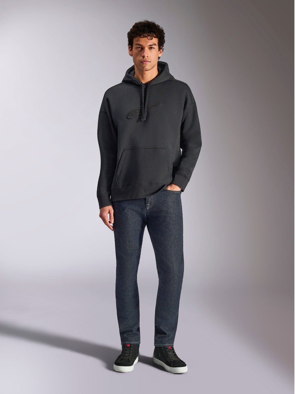 Alpinestars Emergent Oversized Hoodie, casual lifestyle streetwear, zwart met tonaal 3D-geborduurd logo, capuchon met trekkoord, kangoeroezak, oversized pasvorm, gedragen door een model met donkerblauwe jeans en zwarte sneakers.