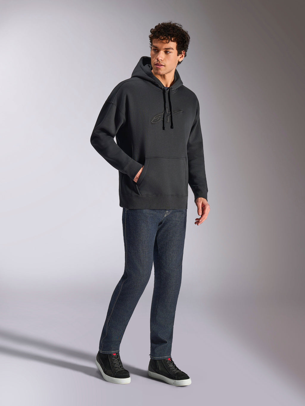Alpinestars Emergent Oversized Hoodie, zwarte casual streetwear, ton-sur-ton 3D geborduurd Astars-logo op de borst, kangoeroezak, oversized pasvorm, gedragen door een mannelijk model met donkerblauwe denim jeans en zwarte sneakers