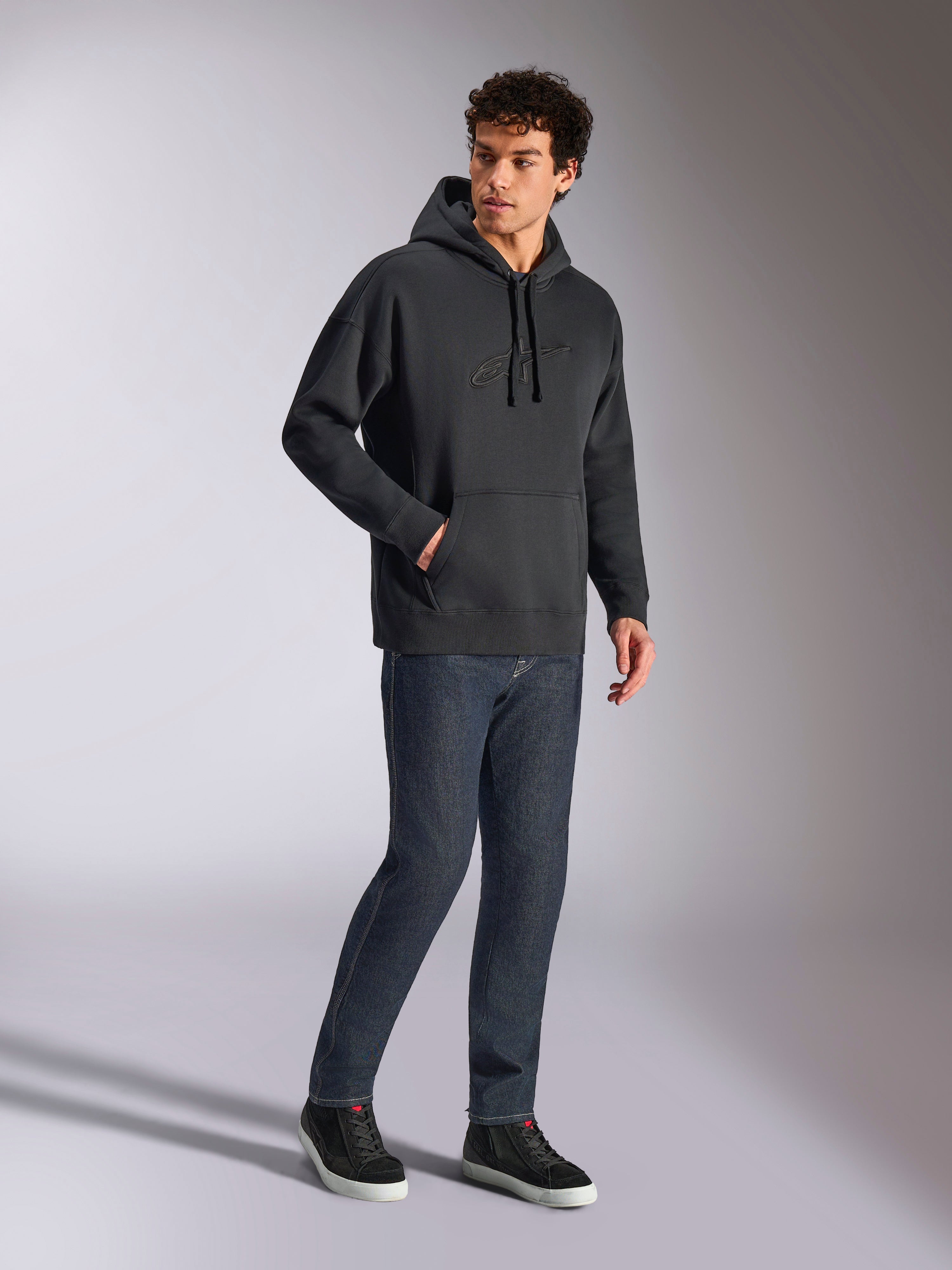 Alpinestars Emergent Oversized Hoodie, zwarte casual streetwear, ton-sur-ton 3D geborduurd Astars-logo op de borst, kangoeroezak, oversized pasvorm, gedragen door een mannelijk model met donkerblauwe denim jeans en zwarte sneakers