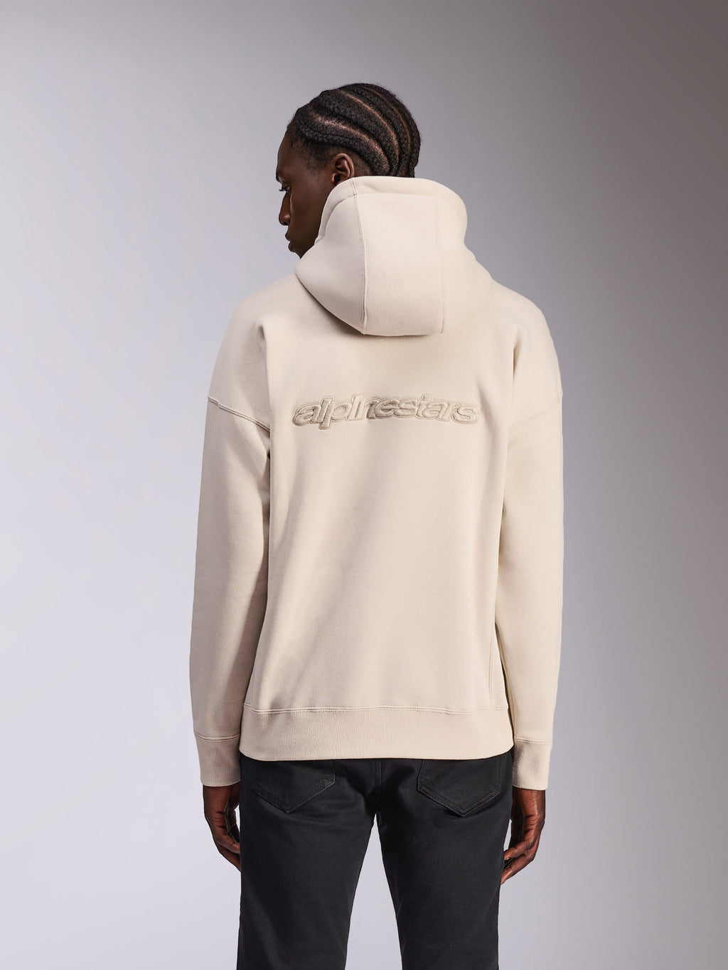 Alpinestars Emergent Oversized Hoodie, casual streetwear, zandkleurige off-white, met 3D-geborduurd Alpinestars-woordlogo op de rug, oversized pasvorm met geribbelde zoom en manchetten, achteraanzicht op model met zwarte broek