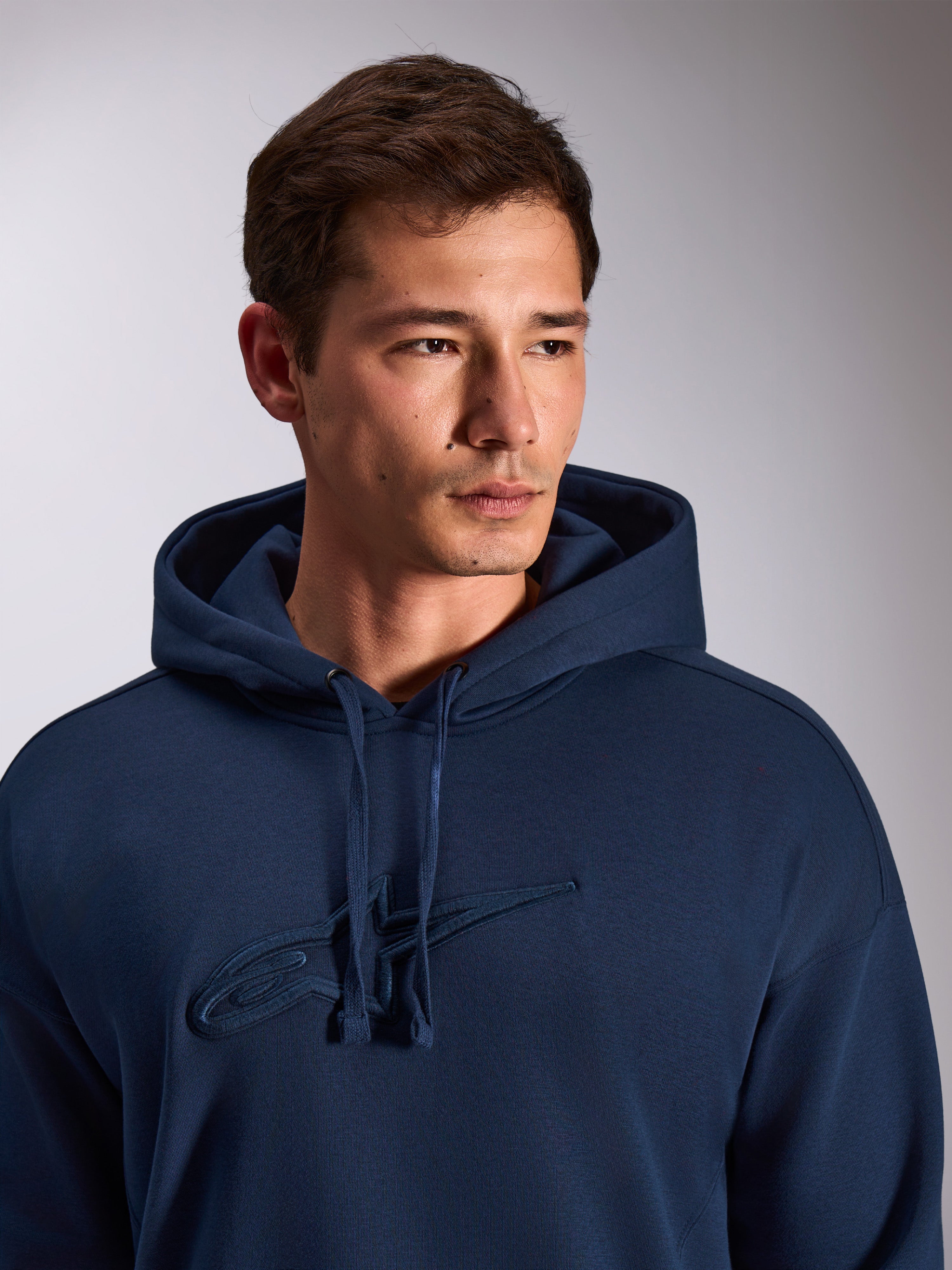 Alpinestars Emergent Oversized Hoodie, marineblauw, close-up van model in pullover fleece hoodie, voorzien van tonaal 3D-geborduurd Astars-logo op de borst, verstelbare capuchon met trekkoord, casual motorsport streetwear