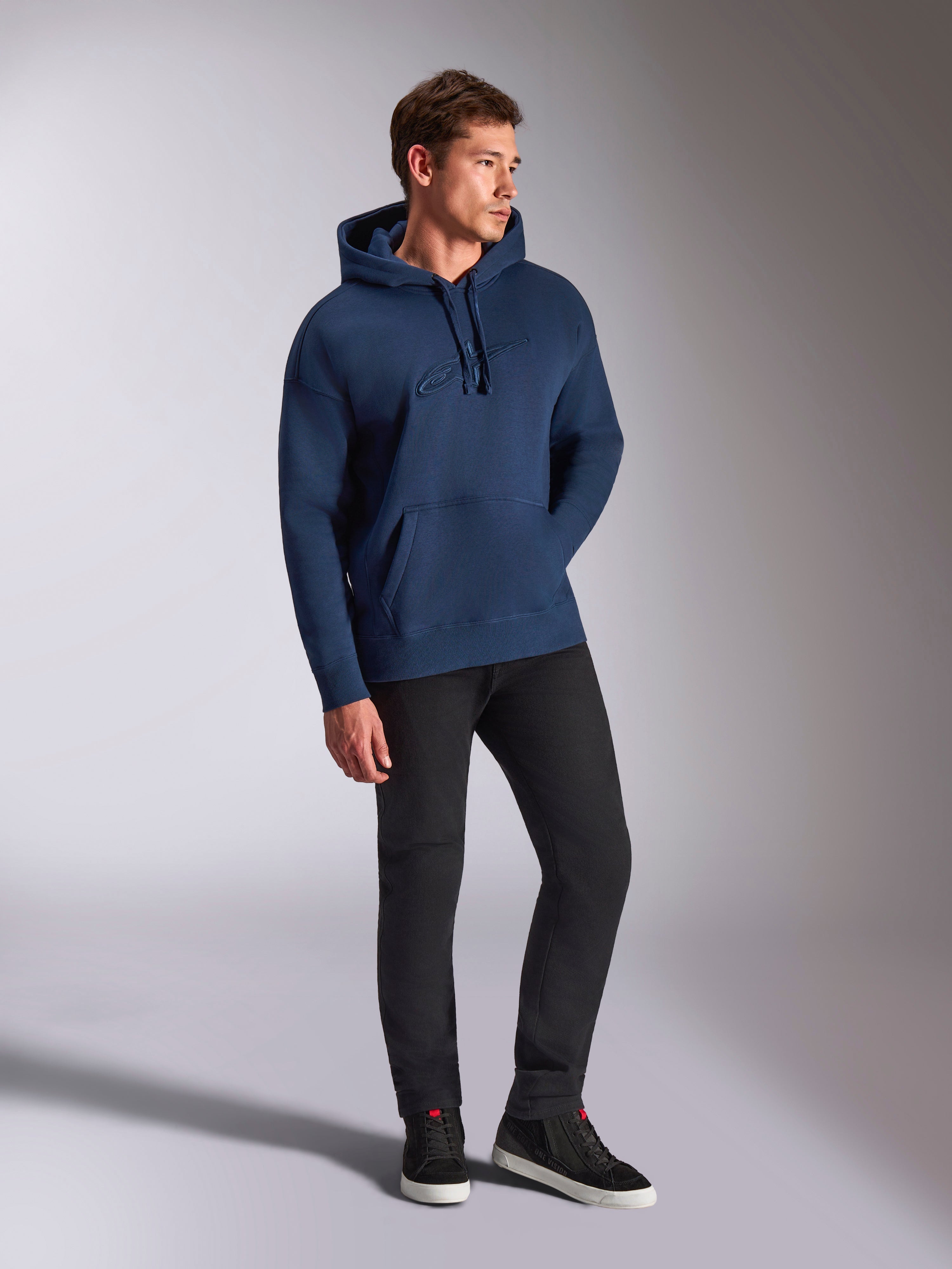 Alpinestars Emergent Oversized Hoodie, navy blauw, gedragen door een mannelijk model met zwarte broek en sneakers, voorzien van een 3D-geborduurd Astars-logo, kangoeroezak en capuchon met trekkoord, casual motorsport-geïnspireerde streetwear.